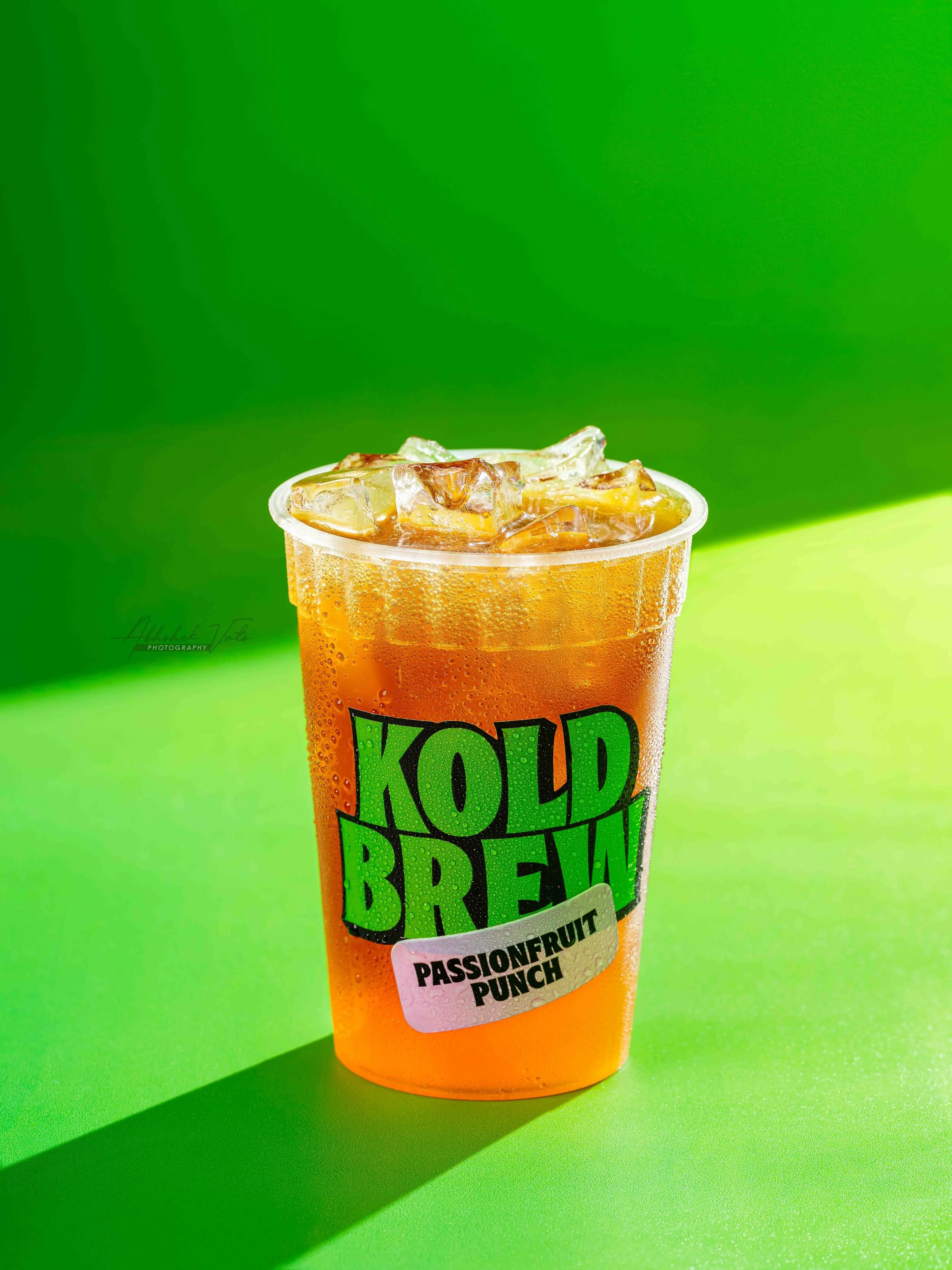 A cold beverage in a plastic cup with ice, labeled as 'KOLD BREW,' flavor 'Passionfruit Punch,' with a bright green background and shadow.
