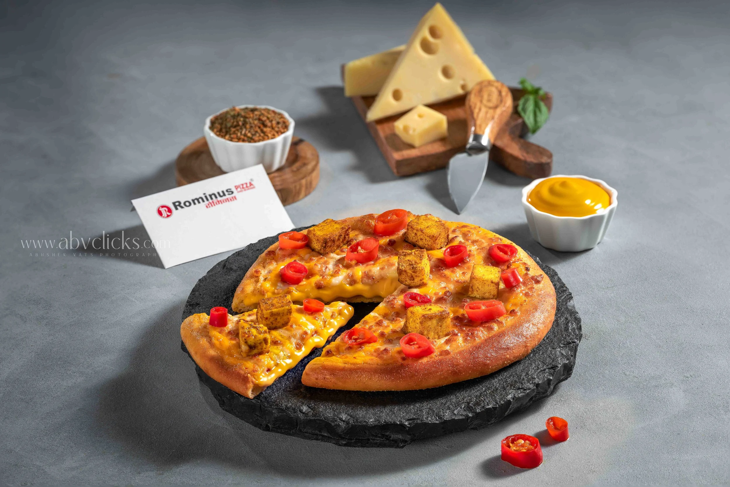 Close-up of a pizza with cheese, chopped tomatoes, and tofu cubes on a round black stone slab. In the background, there are slices of Swiss cheese on a wooden board, a small bowl of ground spices, and a dish of honey mustard sauce on a gray surface.