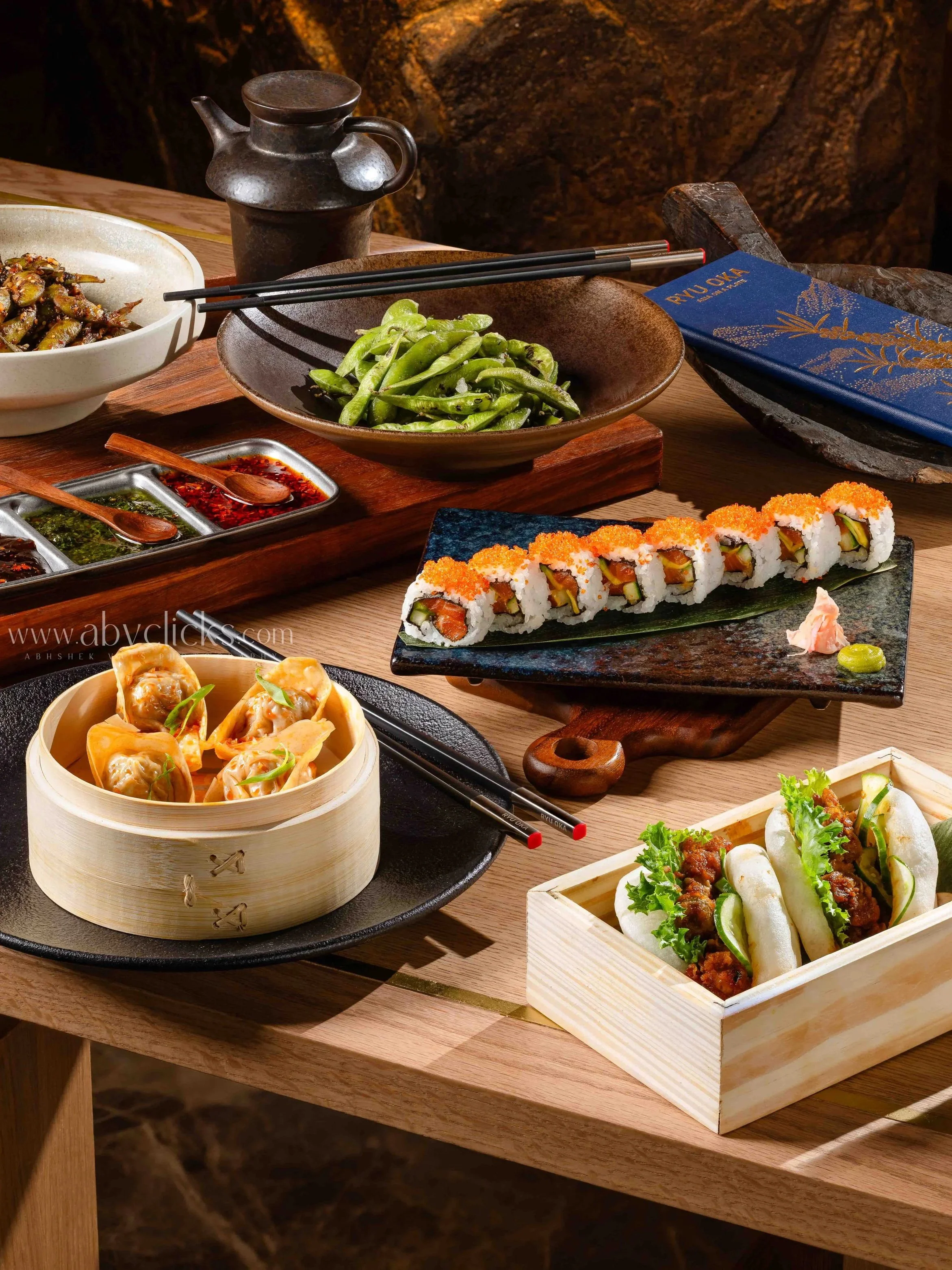 Various Asian dishes on a wooden table, including sushi rolls with orange fish roe, dim sum dumplings in a bamboo steamer, and plates of cooked vegetables and sauces.