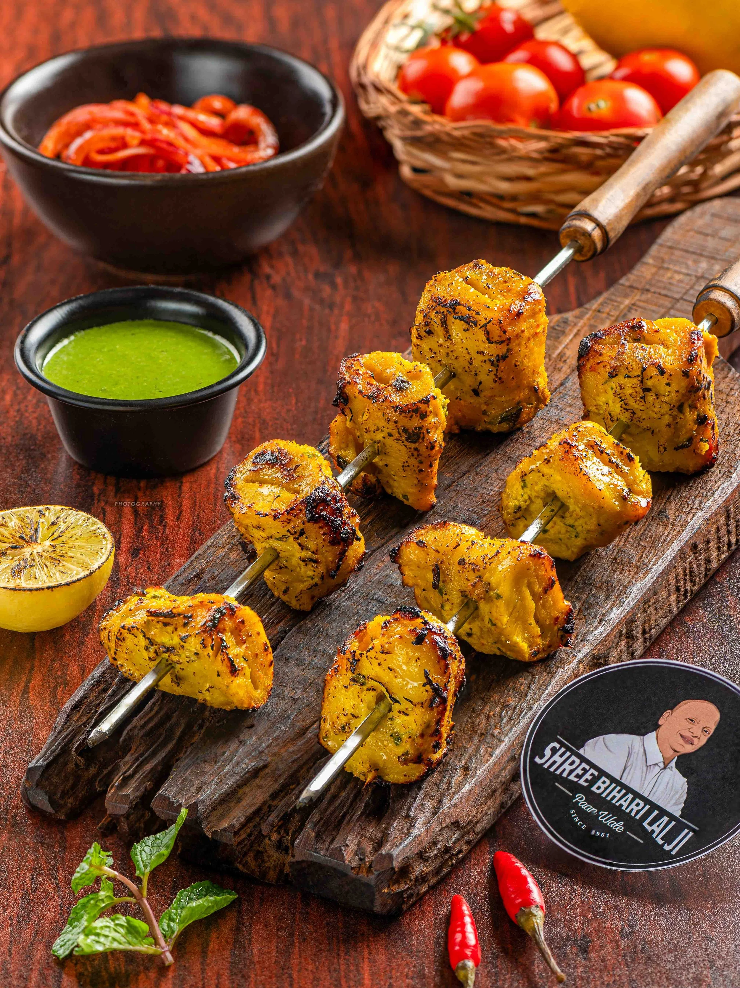 Close-up of grilled chicken tikka kebabs on a wooden serving board, with a bowl of green chutney, a bowl of tomato-based sauce, cherry tomatoes in a basket, a lemon half, and red chilies on a dark wooden surface.