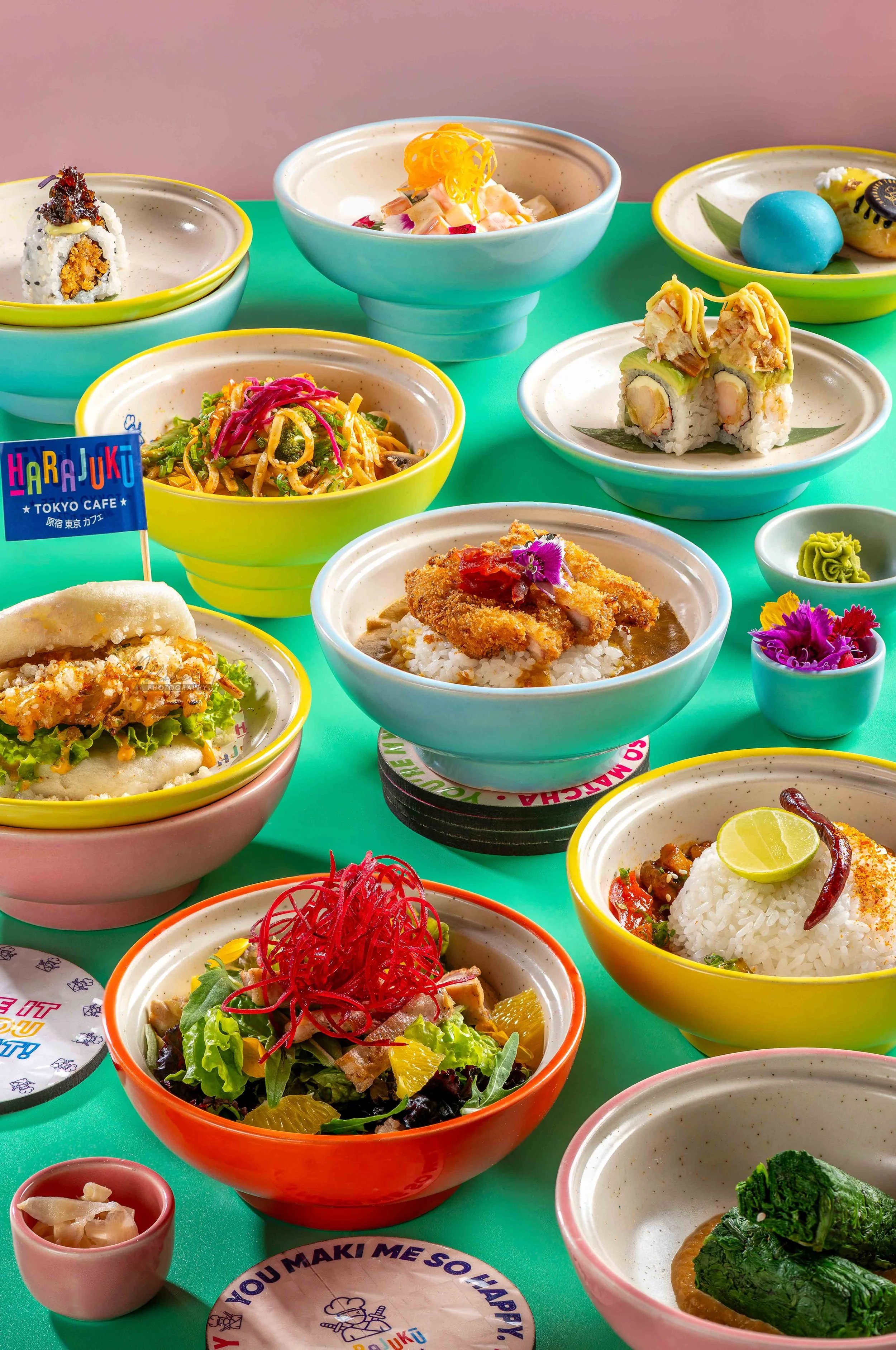 Assorted colorful Japanese dishes and side dishes on a green table, including sushi rolls, salads, rice bowls, and desserts.