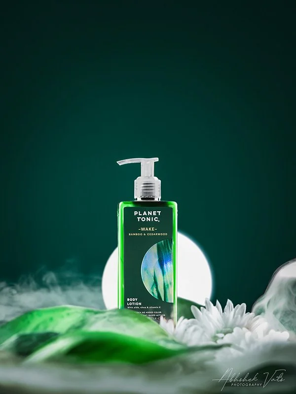 A green bottle of Planet Tonic Wake body lotion on a white surface with white flowers in front, green leaves at the bottom, and a bright light behind the bottle creating a glowing effect.