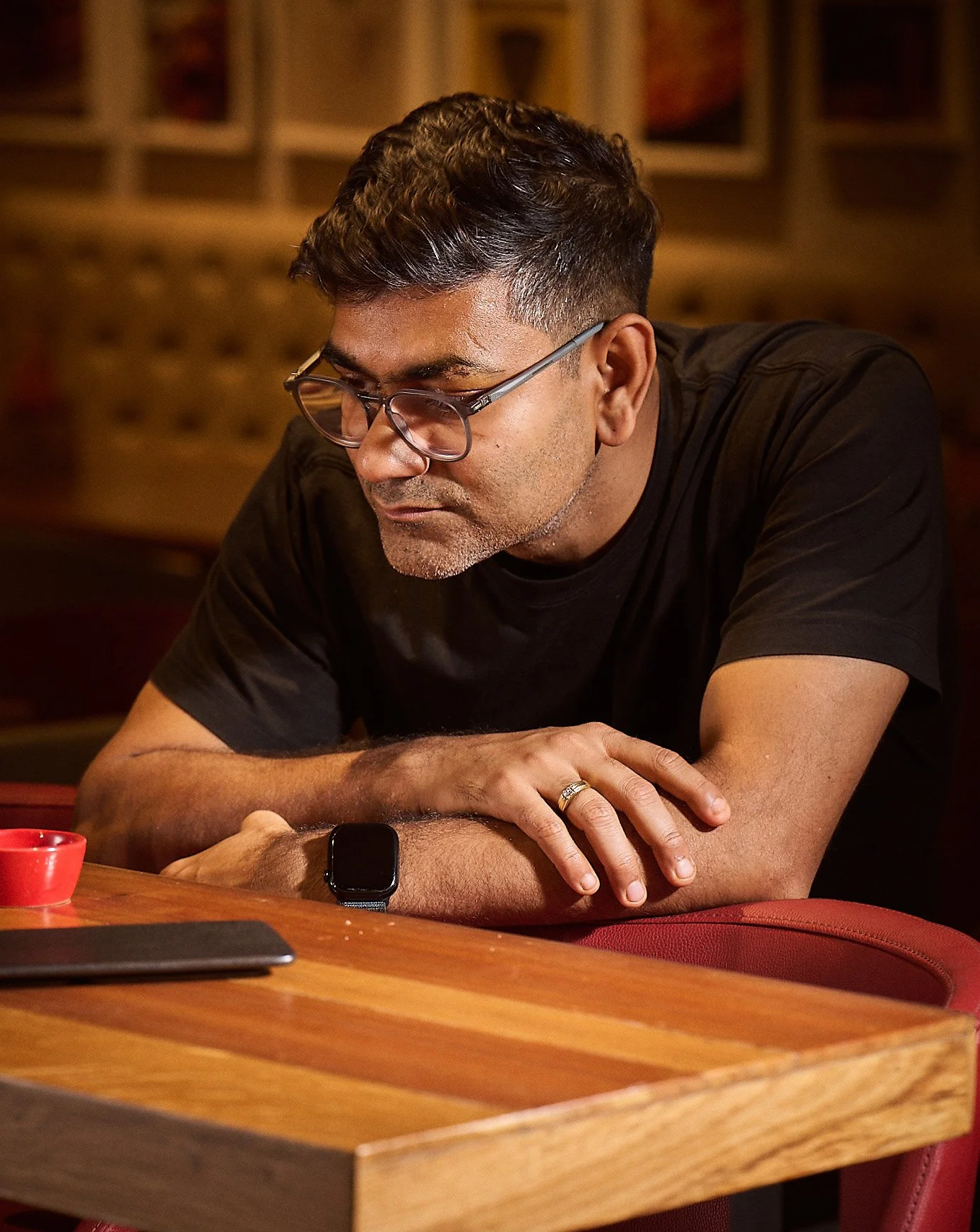 A man with glasses and a watch is sitting at a wooden table, looking down at his phone with his arms crossed on the table. There is a red cup and a tablet on the table. The background features brown upholstered walls and framed pictures.
