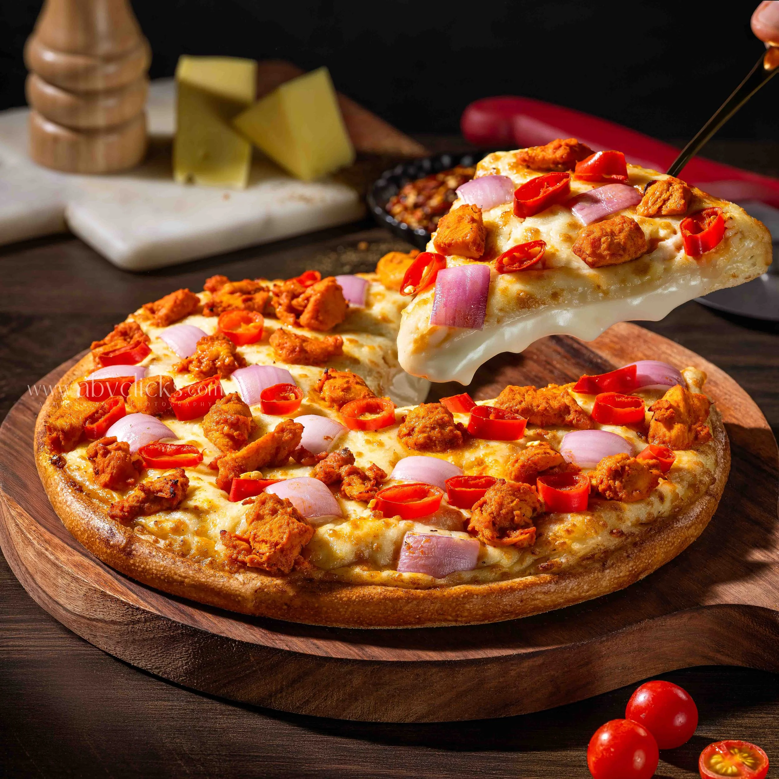 Close-up of a pepperoni pizza slice topped with red chili peppers, jalapenos, and onion, with a wooden pizza board underneath. In the background, there is a cutting board with cheese, red pepper flakes, and a wooden grinder.
