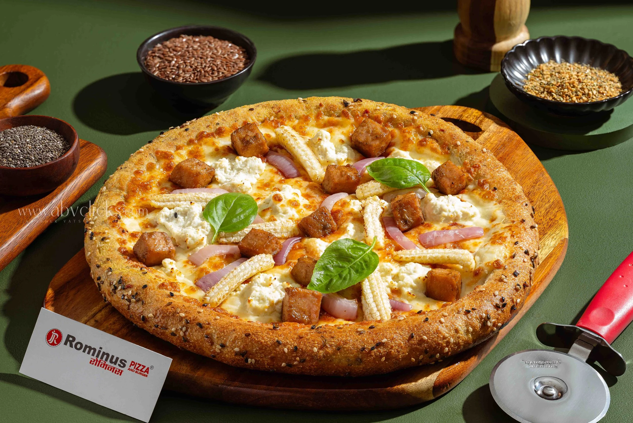 A pizza with a thick crust topped with cheese, fried tofu cubes, sausage slices, and fresh basil leaves, on a wooden serving board. Surrounding bowls contain seeds and grains, a pizza restaurant flyer is partially visible, and a soda can with a red s
