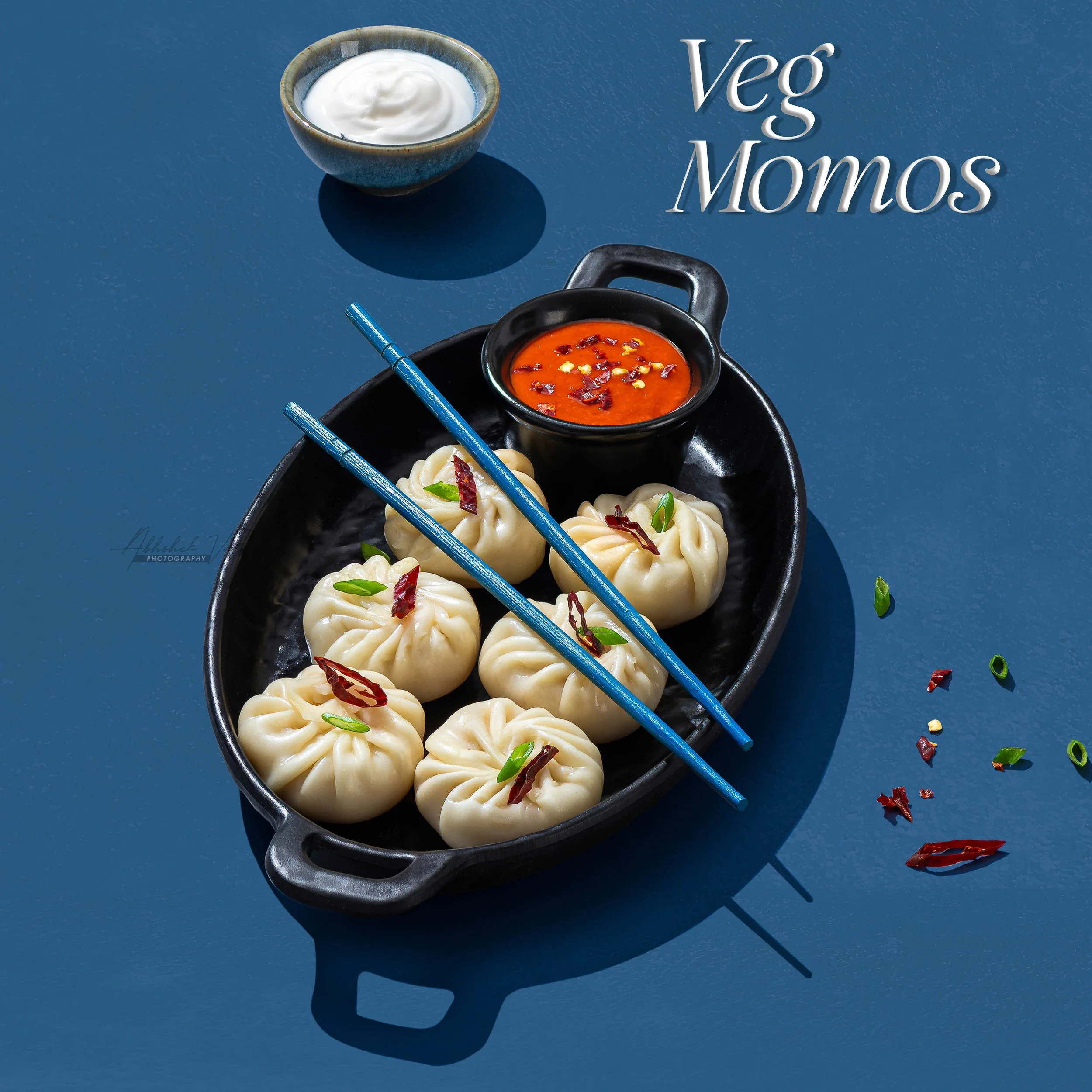 Plate containing steamed dumplings garnished with sliced green onions and red chili flakes, with a small bowl of spicy red dipping sauce in the center, placed on a dark tray. Chopsticks rest on the tray. The background is a blue surface with a shadow