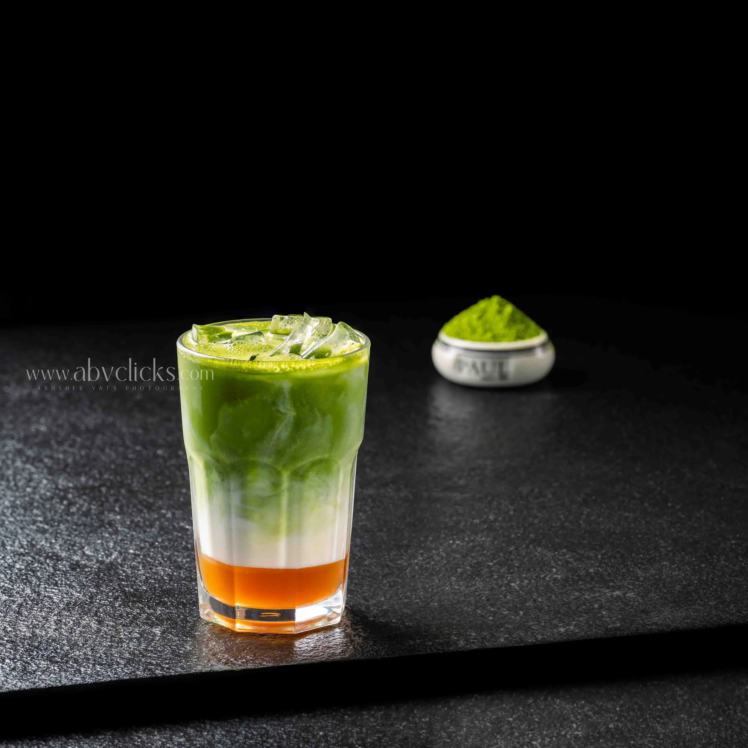 Layered cocktail in a tall glass with an orange base, green middle layer, and topped with ice, lime juice, and ice cubes against a black background.
