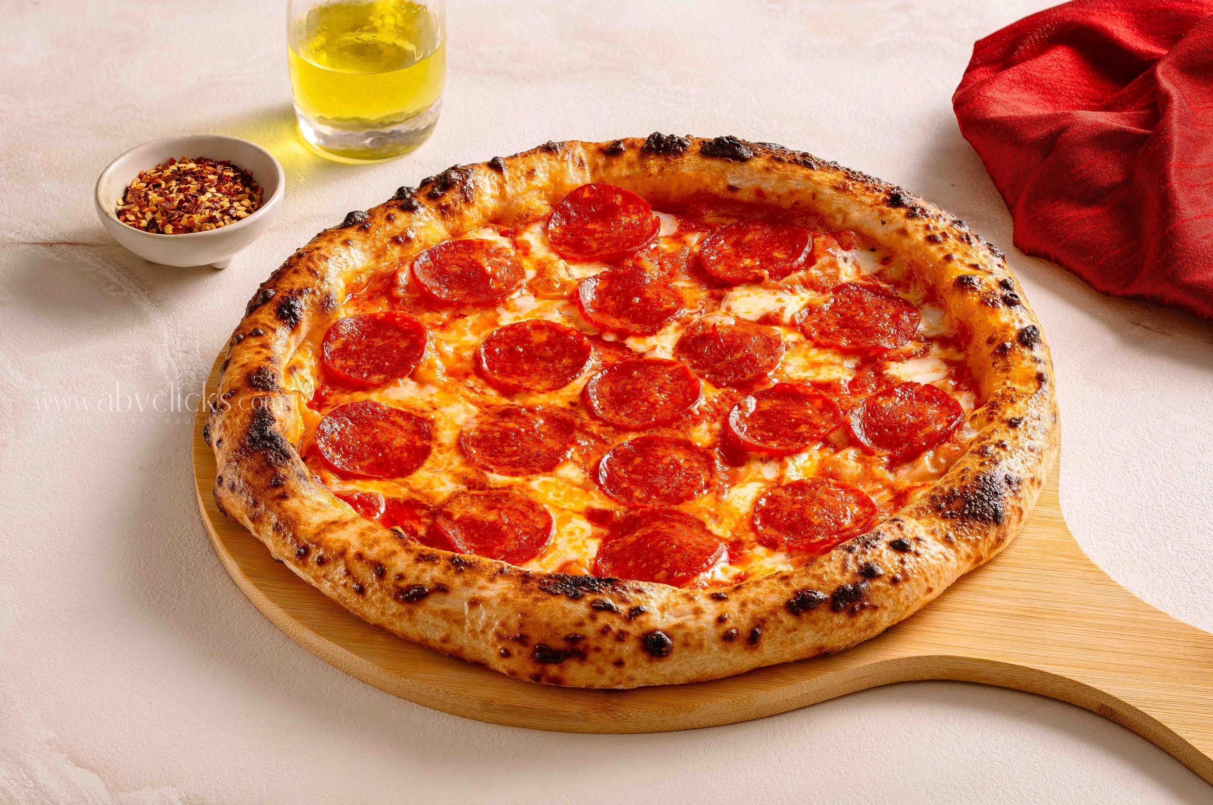 A pepperoni pizza on a round wooden board, with a glass of yellow beverage, a small bowl of red pepper flakes, and a red cloth napkin on a light-colored table.