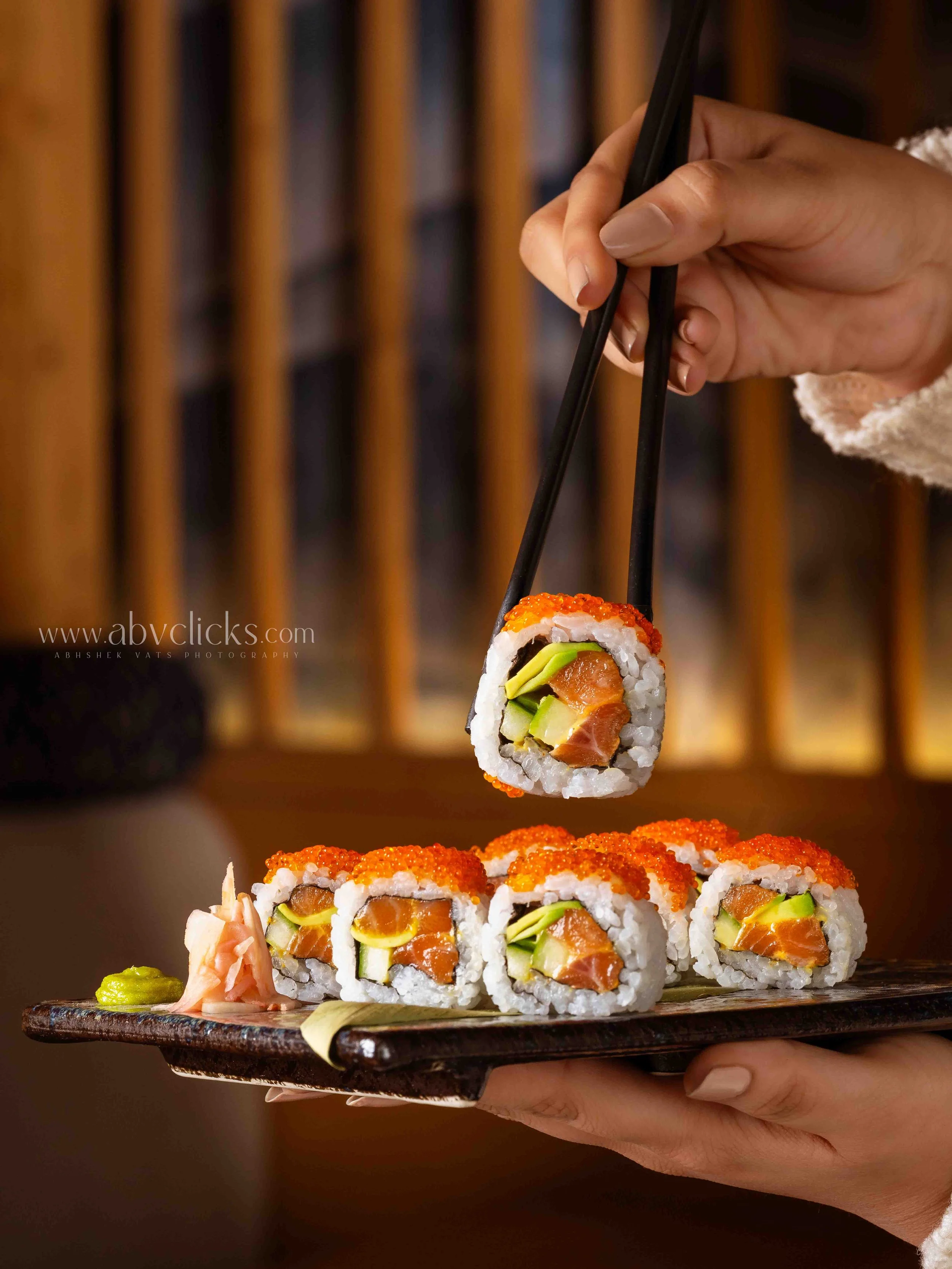 Hand holding a black chopstick with a sushi roll over a dark wooden plate, with more sushi rolls and condiments on a plate in the background.