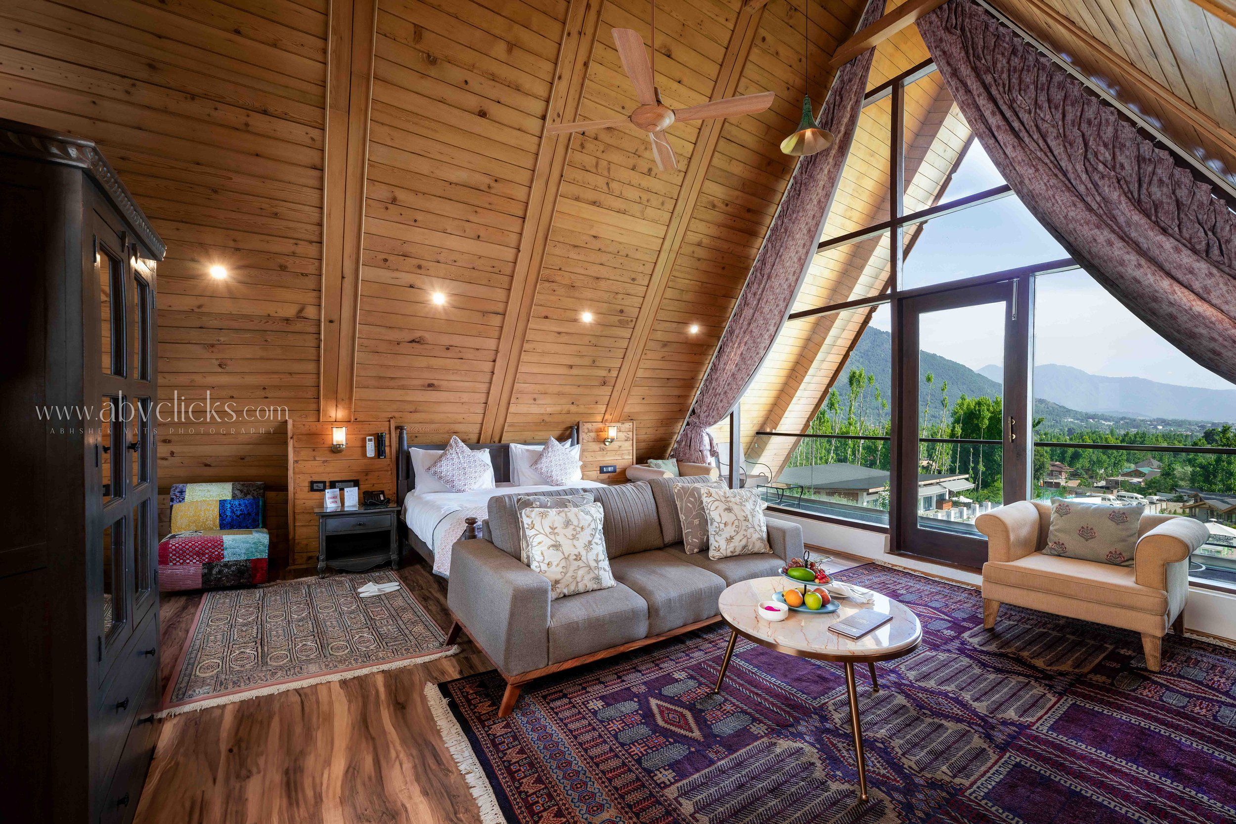 Interior of a modern hotel room with a sloped wooden ceiling, large windows with views of mountains and trees, a bed with pillows, a sofa, a round coffee table with fruit, a wooden ceiling fan, and eclectic furniture and decor.