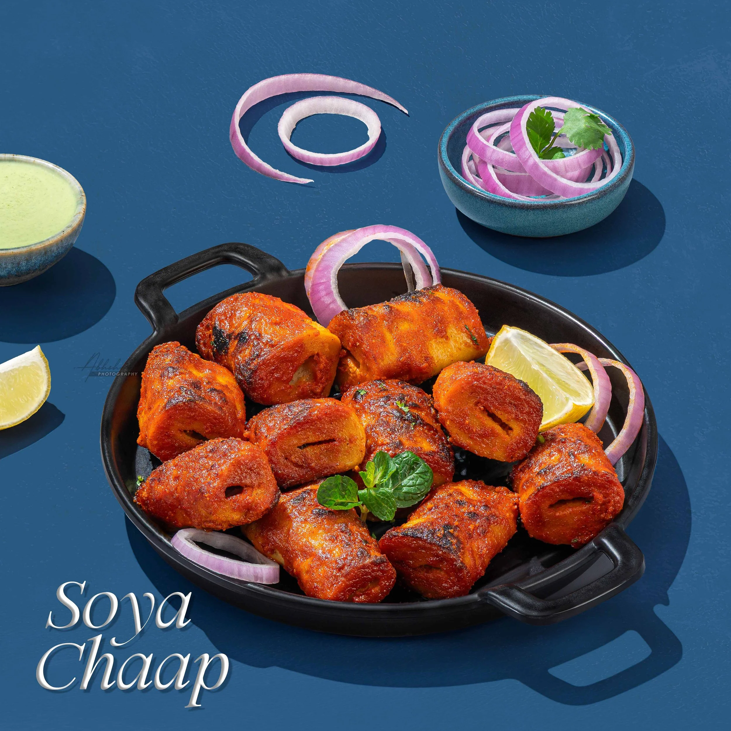 Close-up of a black skillet filled with fried chicken pieces garnished with lemon wedges and herbs, on a blue surface. Surrounding the skillet are sliced red onions, lemon wedges, and a blue bowl with onion rings and cilantro leaves, with part of a g