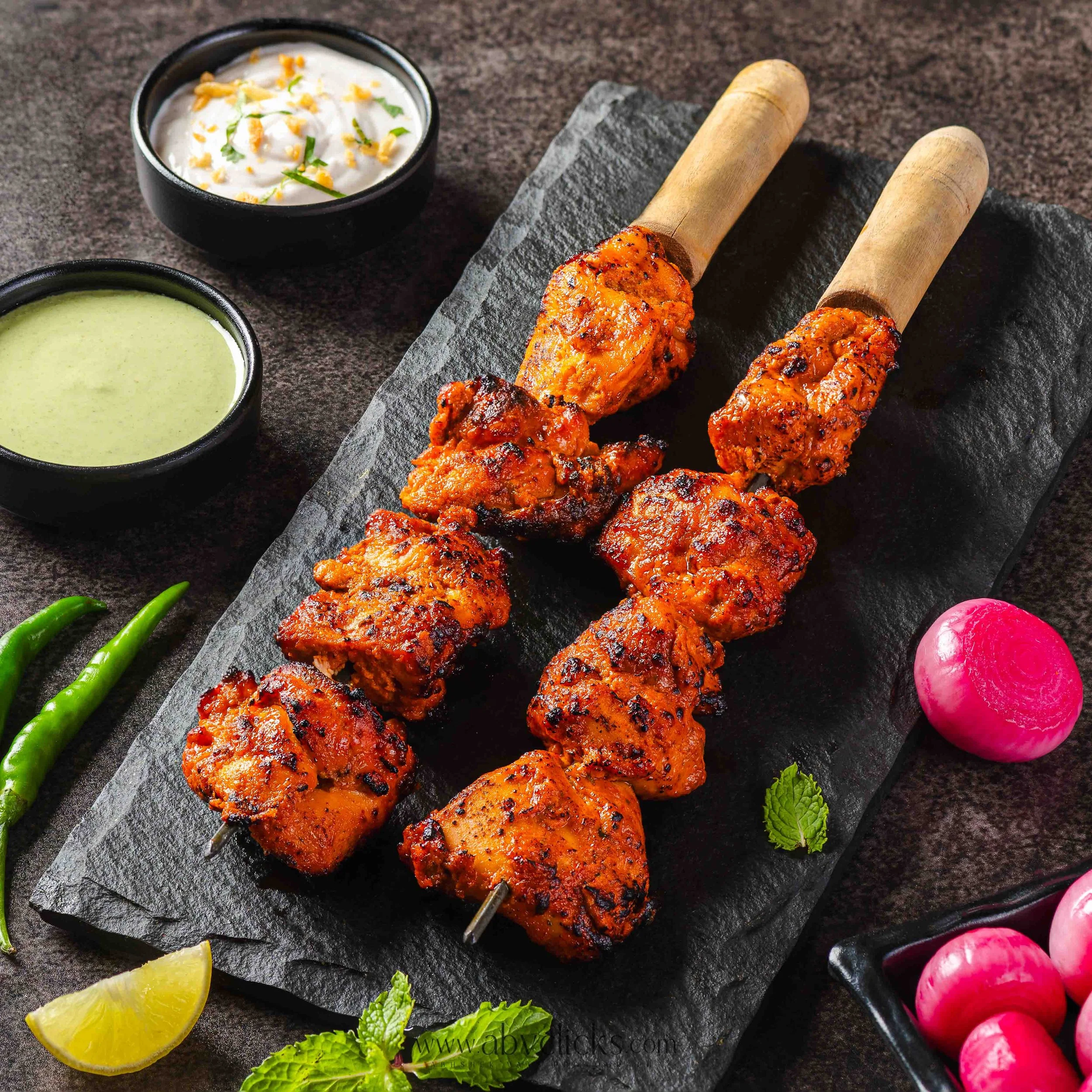 Close-up of skewered tandoori chicken with two small bowls of green and white sauces, lemon wedges, and garnishes on a black slate serving platter.