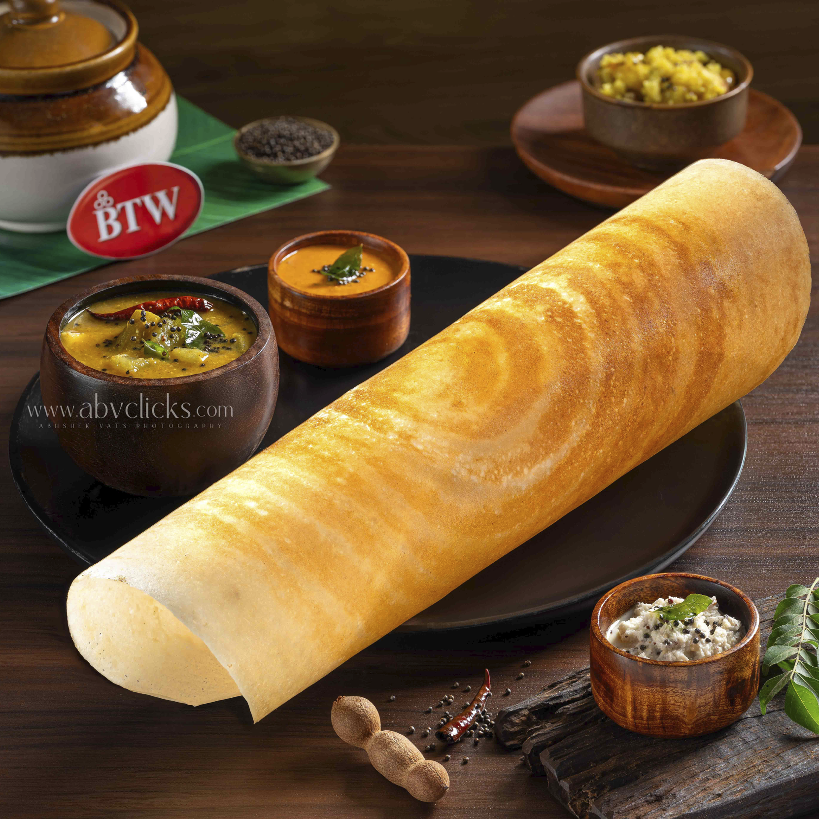 A large rolled Indian roti with small bowls of various Indian dishes, spices, herbs, and condiments on a wooden table.