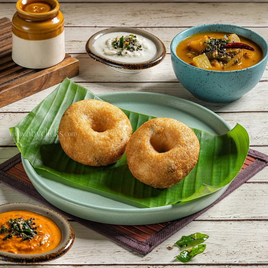 Two deep-fried doughnuts on a banana leaf on a green plate, with dipping sauces and curry in separate bowls around it.