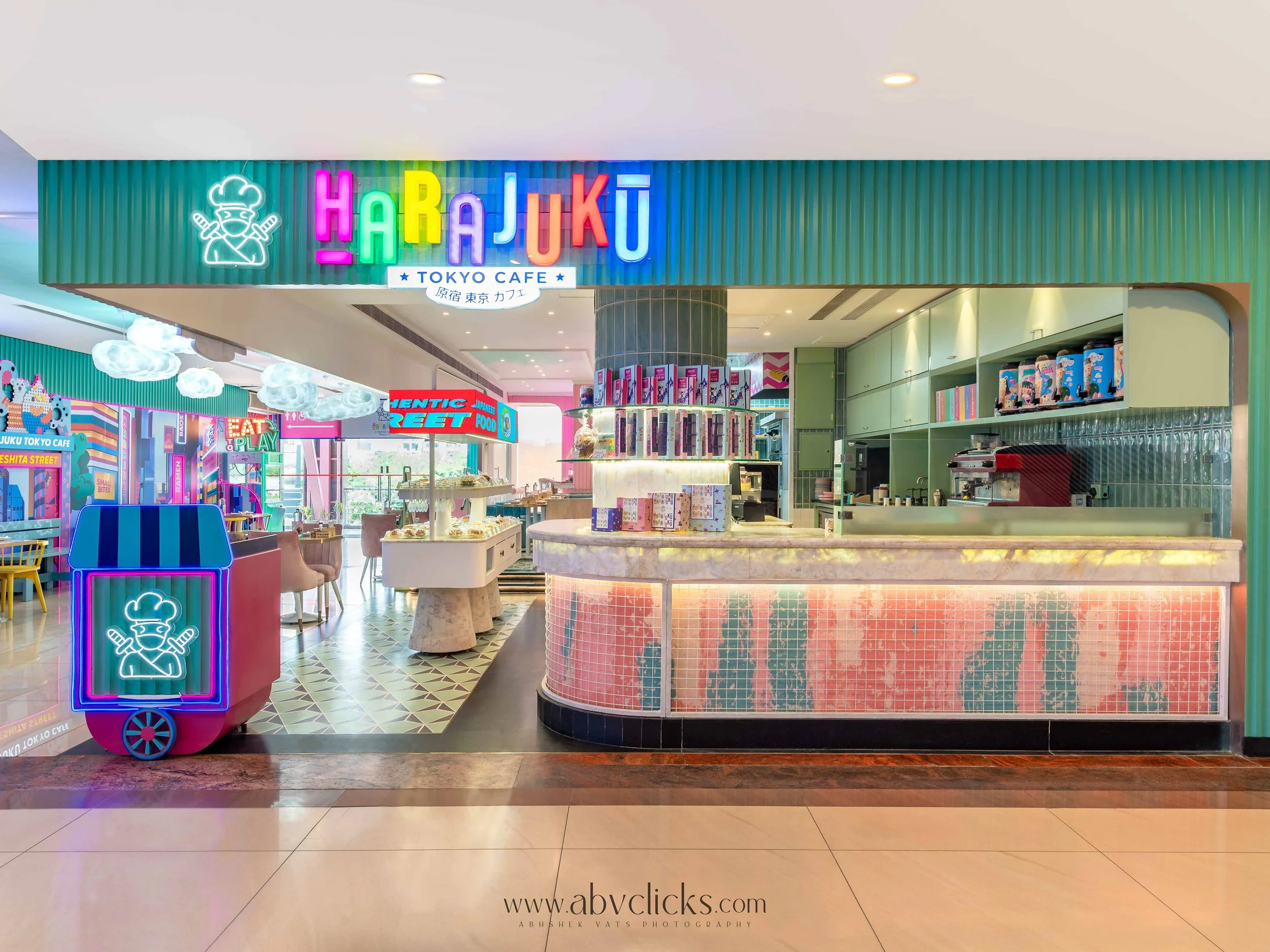 Neon sign reading 'Harajuku Tokyo Cafe' with cartoon chef logo, pastel-colored interior with tables, chairs, and decorative elements.