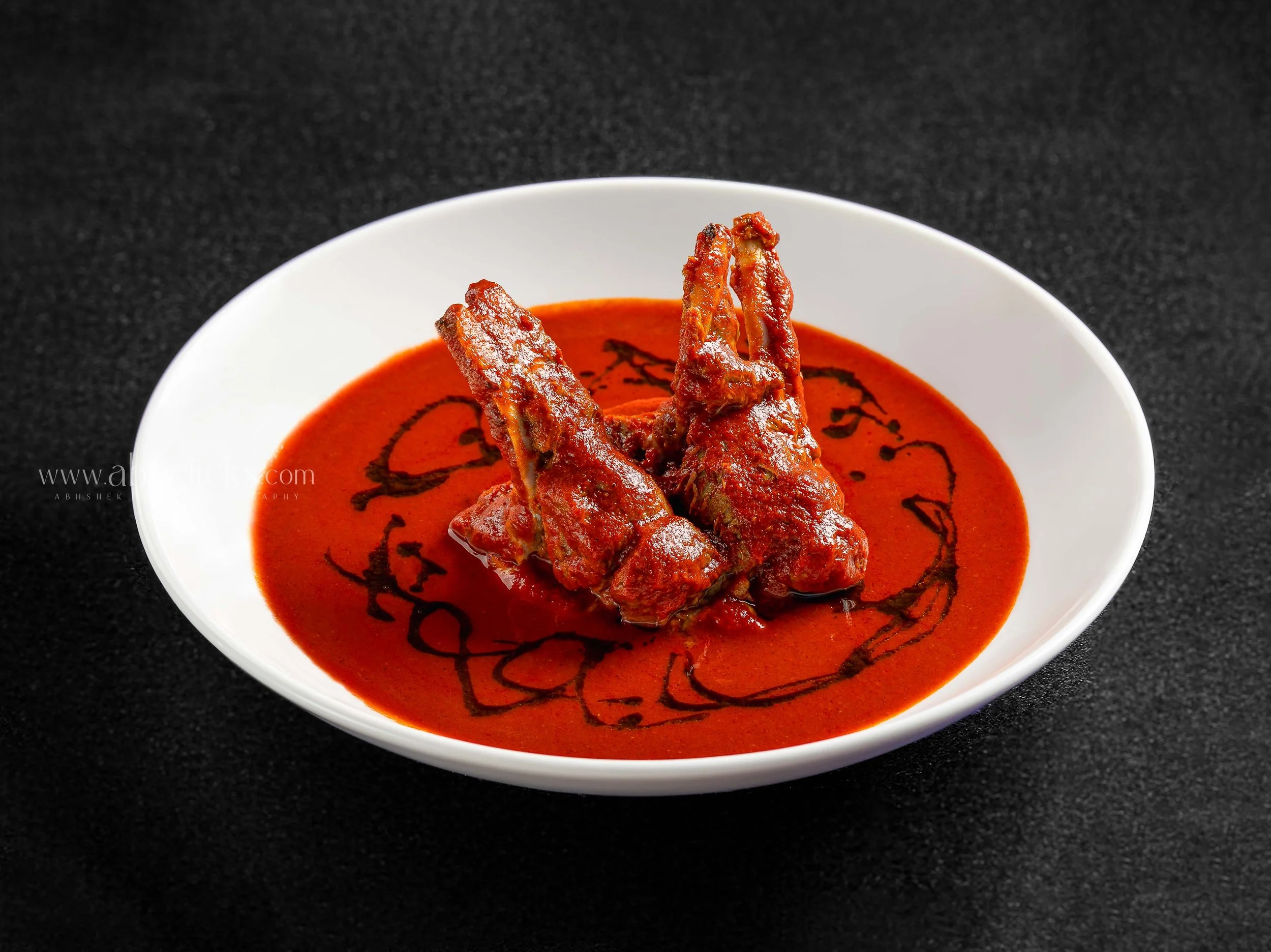A white bowl contains a red curry with two pieces of meat and a dark drizzle on top, placed on a black surface.