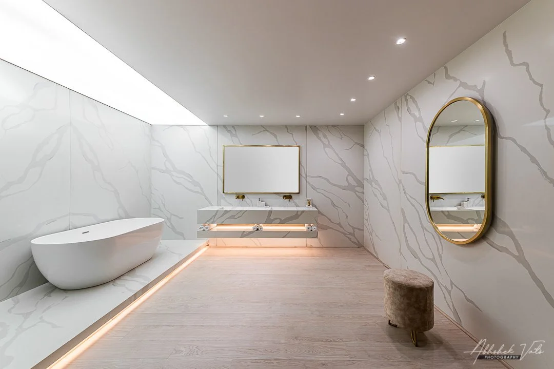 Modern, minimalist bathroom with white marble walls, a free-standing bathtub, a floating vanity with a large mirror, a round mirror on the right wall, and a small beige stool.