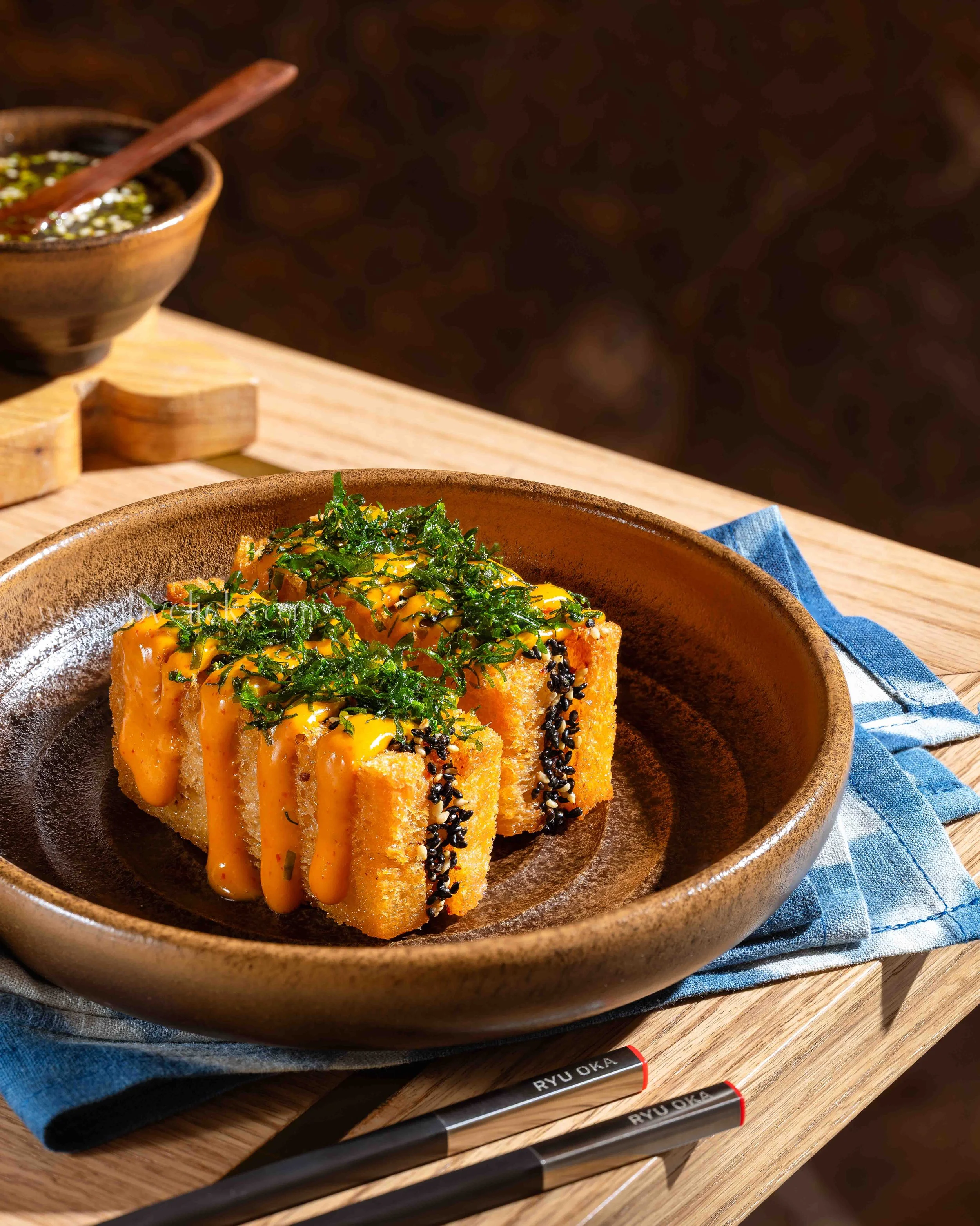 A bowl of sushi topped with chopped green herbs and drizzled with orange sauce on a wooden table, with a blue napkin underneath and black chopsticks with white writing.