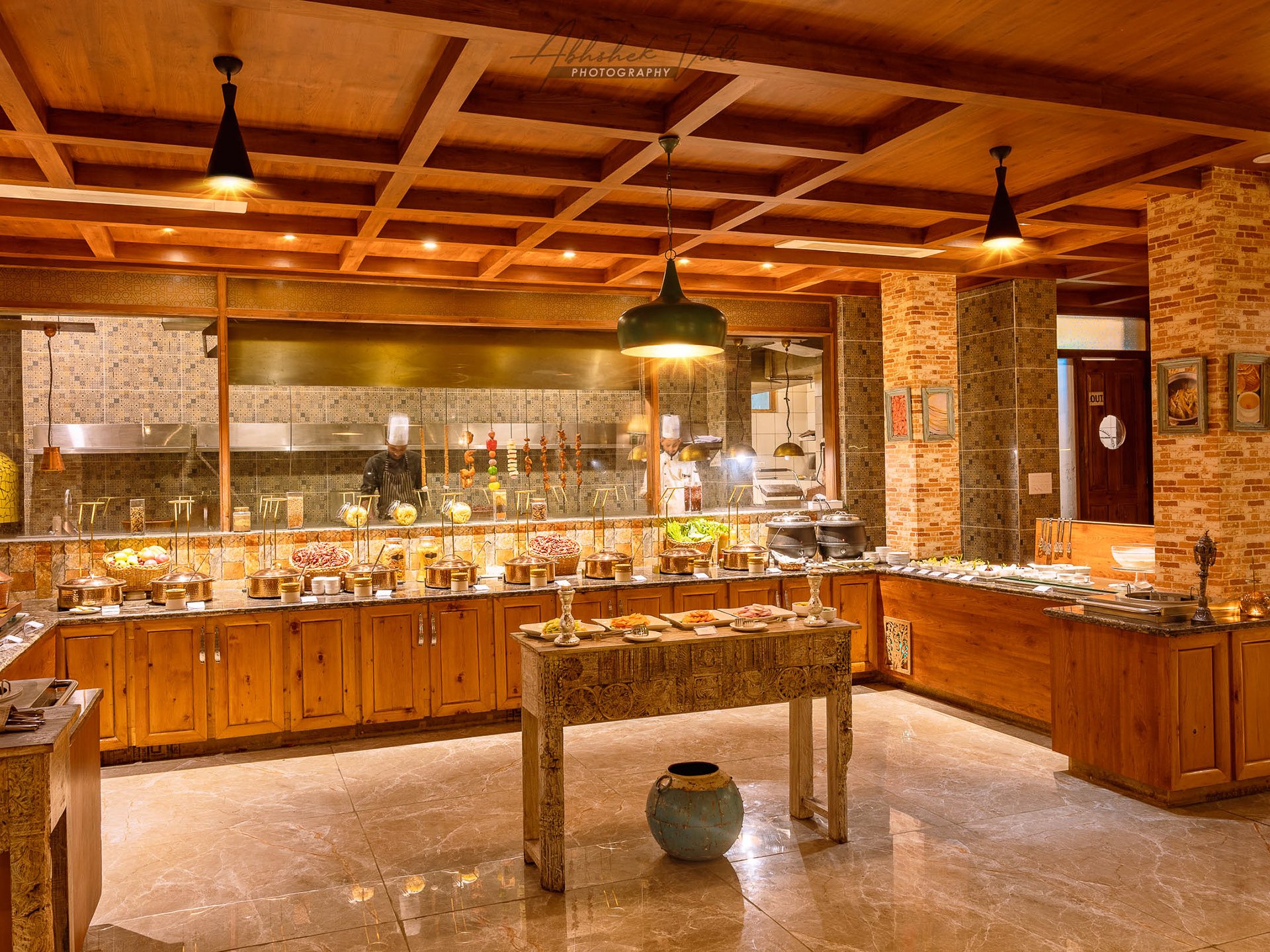 A warm, rustic buffet setup in a restaurant with wooden cabinets, a tiled backsplash, hanging lights, and a marble floor. Chefs are seen behind the counter preparing food.