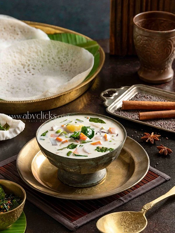A bowl of Indian-style vegetable curry with coconut milk, served on a brass plate, surrounded by papadums, spices, a cup, and a small bowl of salad, with a gold spoon nearby.