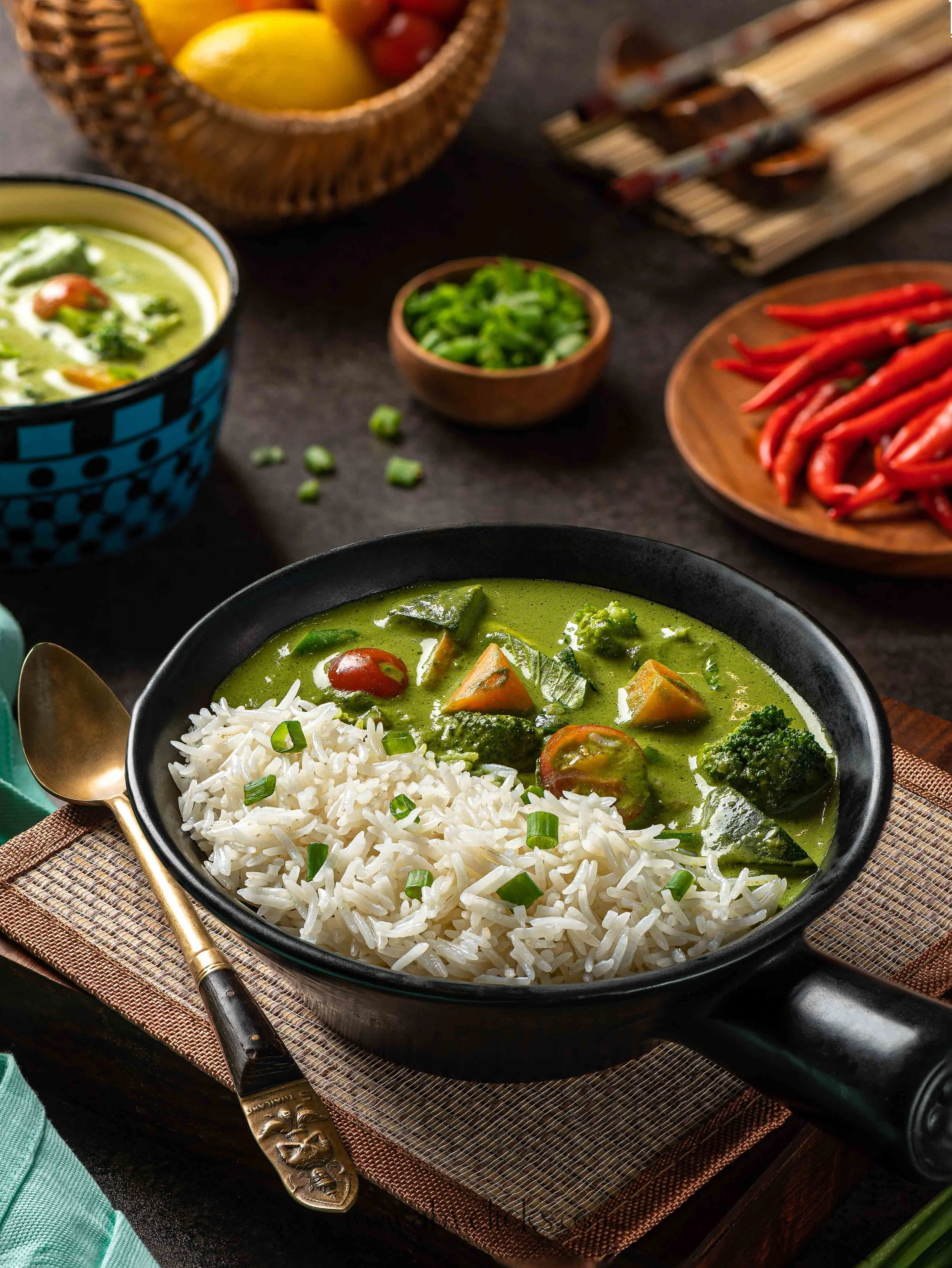 A bowl of green vegetable soup with rice and chopped green onions, surrounded by small bowls of chopped green peppers and red chili peppers, on a woven placemat with a vintage spoon, set on a dark wooden table.