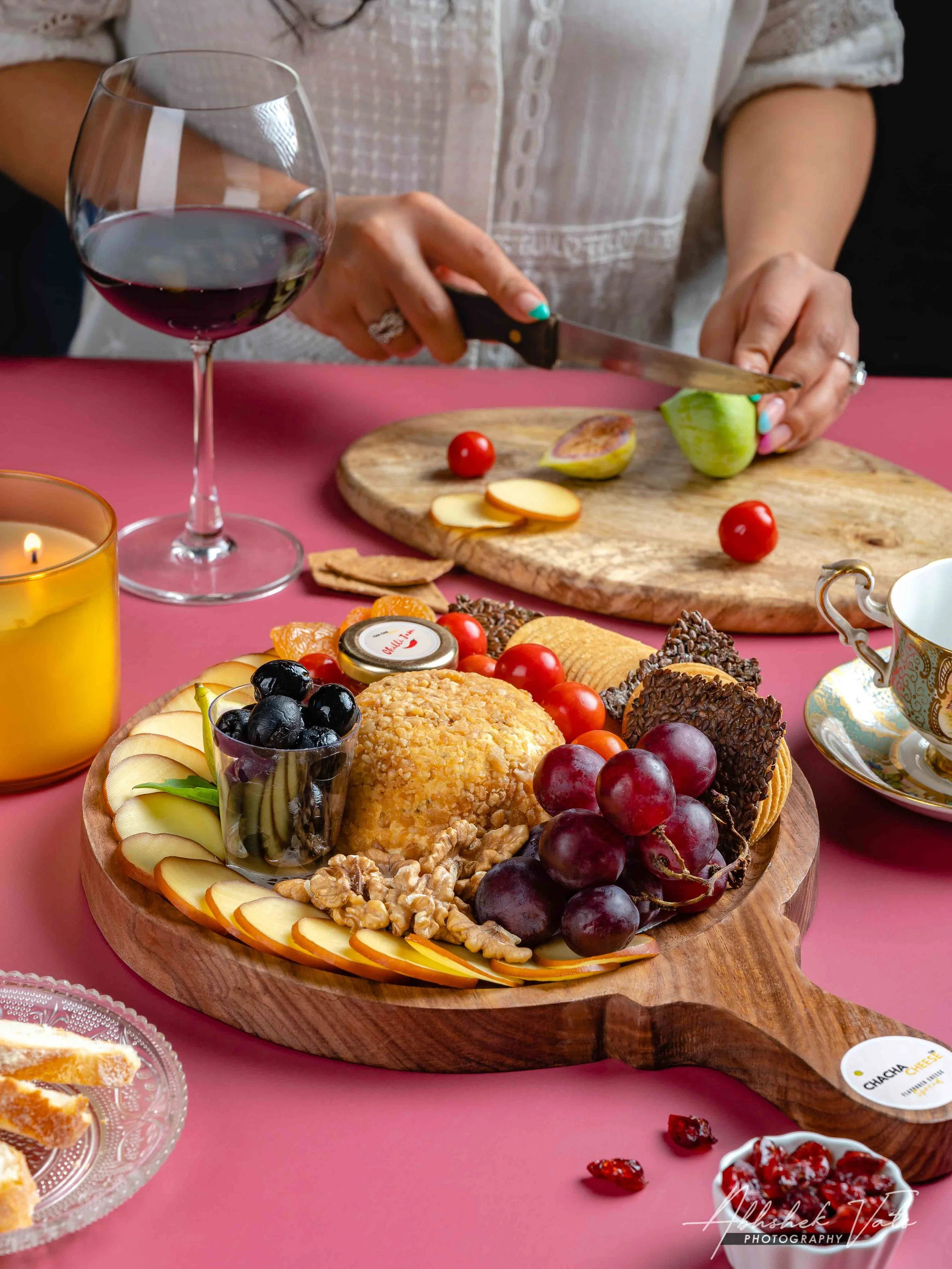 A wooden serving tray on a pink table holds an assortment of cheeses, fresh grapes, cherry tomatoes, walnuts, cookies, and a glass of red wine. In the background, a woman is slicing a fig on a cutting board with other food items and a lit candle near
