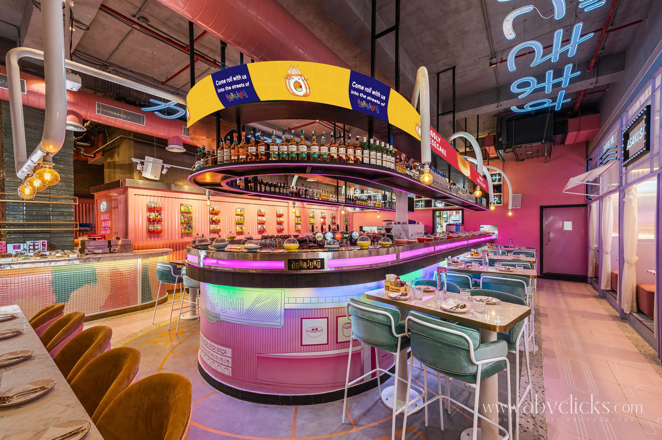 Interior of a vibrant restaurant or cafe with neon pink and blue lighting, circular bar with liquor bottles, and colorful decor, including pink walls and elegant seating.