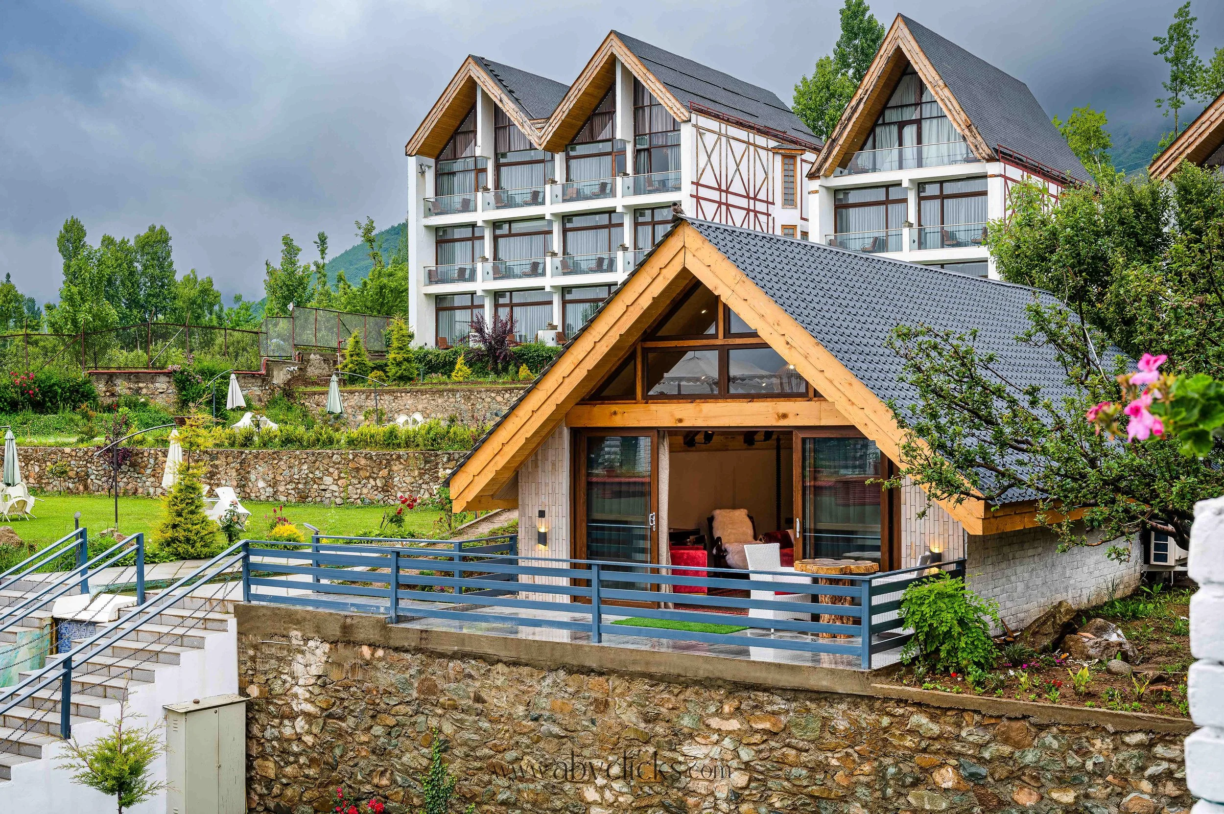 For brand Rahbag by the Orchard, a multi-story building with a mix of modern and traditional architecture, featuring large glass windows, balconies, and a mountain backdrop with trees.