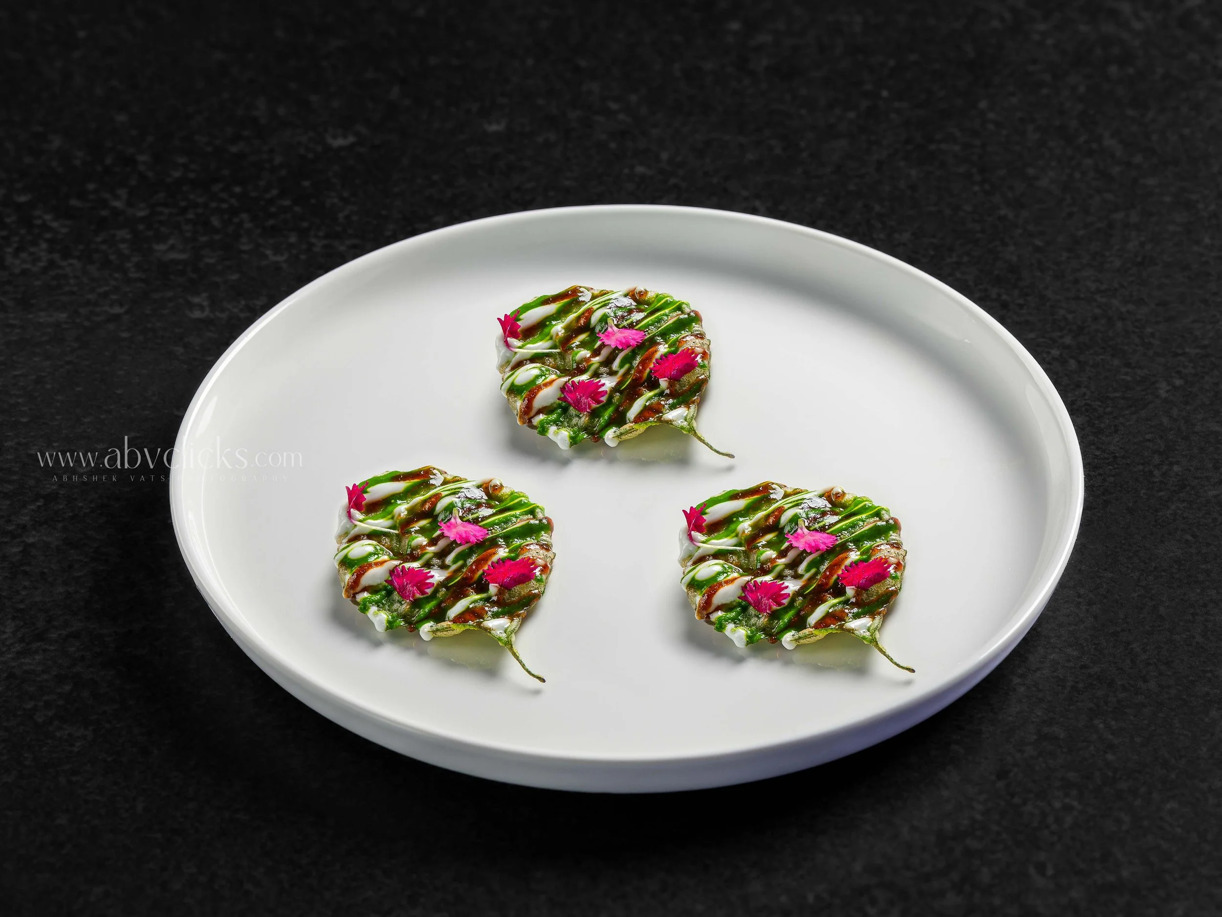 Heart-shaped sushi topped with green sauce, white sauce, and drizzled with brown sauce, garnished with pink edible flowers on a white plate against a black background.