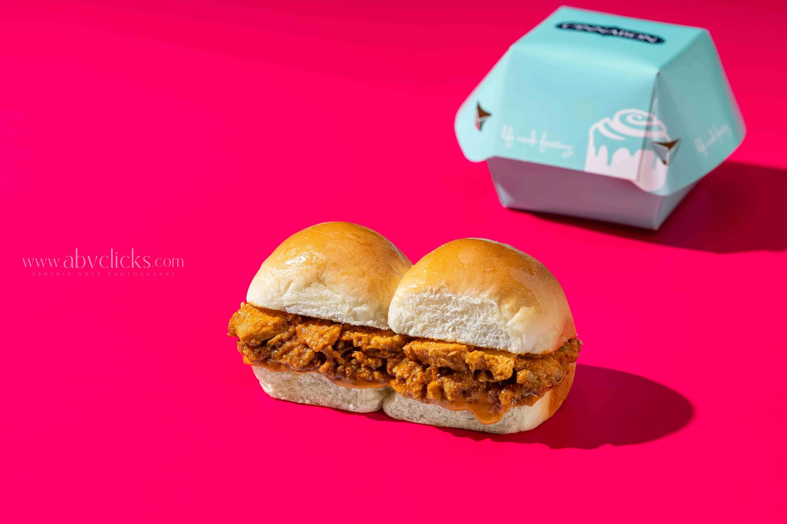 Close-up of a slider burger with a spicy meat filling on a bright pink background, with a small light blue takeout box nearby.