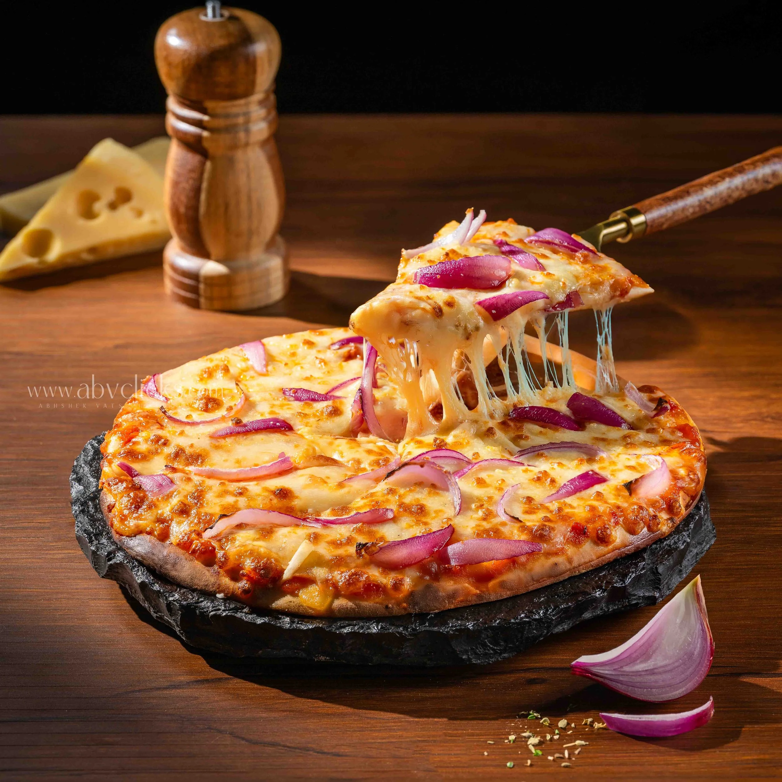 A slice of cheese pizza topped with red onions and melted cheese is being lifted from a pizza on a black stone with cheese stretching as it is pulled away. There is a wooden pepper grinder and a piece of cheese and red onion slices on a wooden table.
