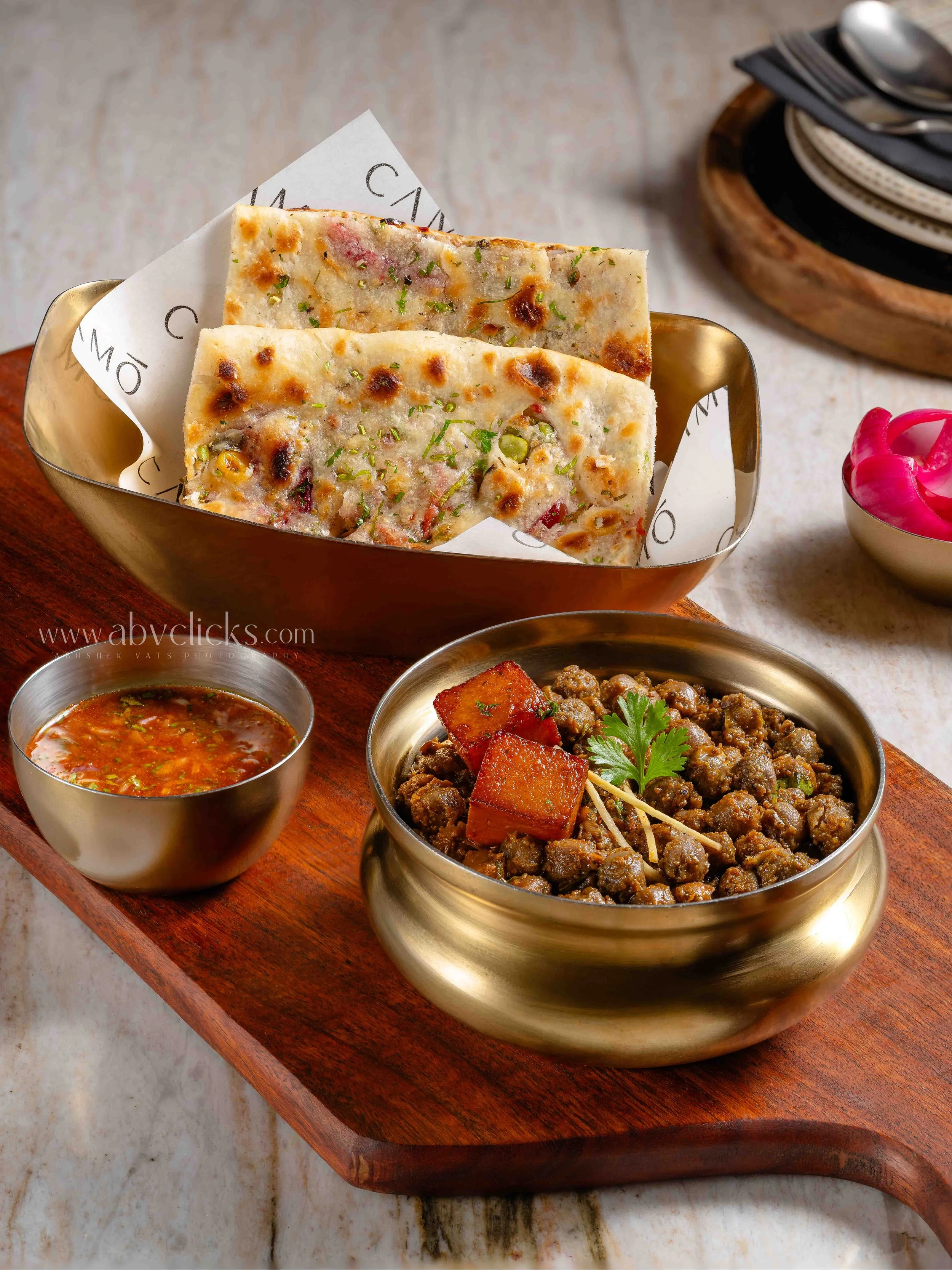 Close-up of Indian food served in metallic bowls on a wooden tray, including a spicy lentil curry, minced meat dish garnished with coriander, and flatbread with herbs, accompanied by small bowls of salsa and pickles on a marble surface.