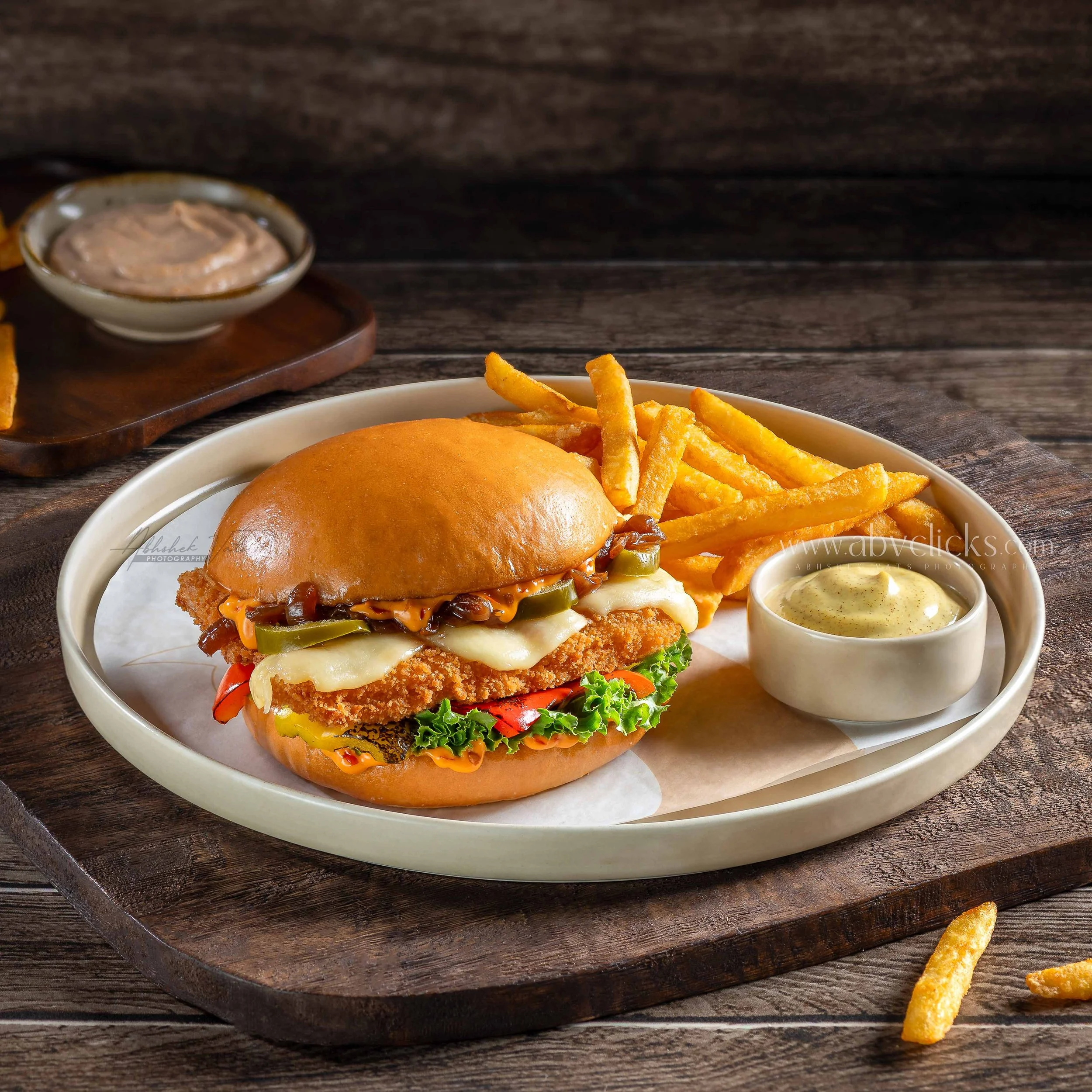 Close-up of a plate with a fried chicken sandwich topped with lettuce, tomato, and pickles, alongside crispy French fries and a small dish of dipping sauce, on a wooden table.