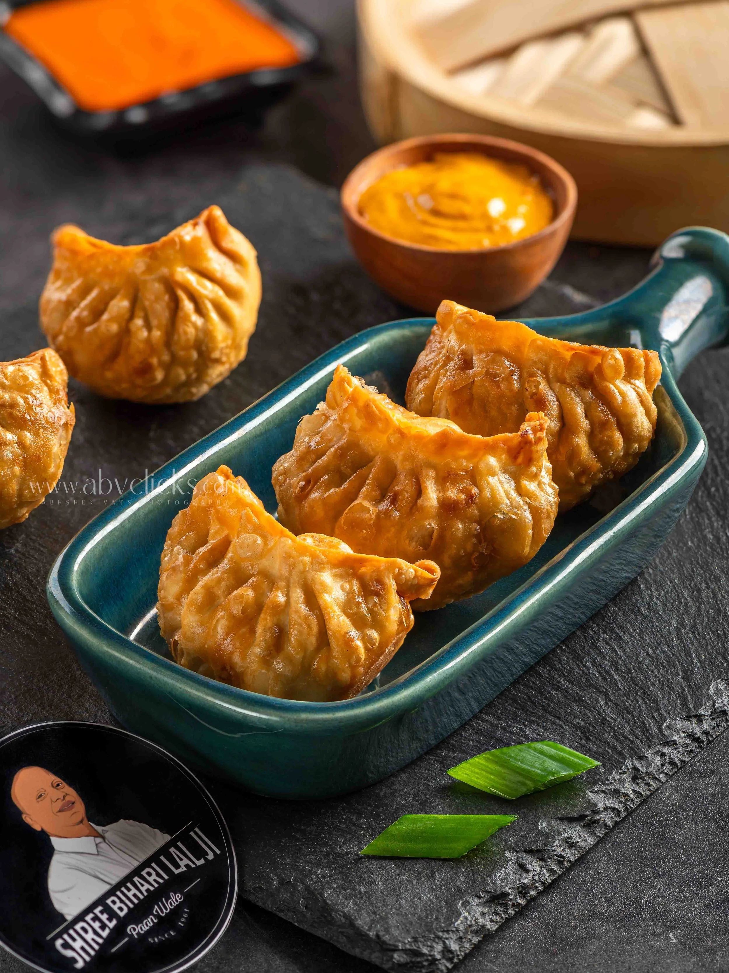 Close-up of several golden fried momos in a teal serving dish, with a small bowl of dipping sauce and a bowl of shredded cabbage in the background on a black surface.