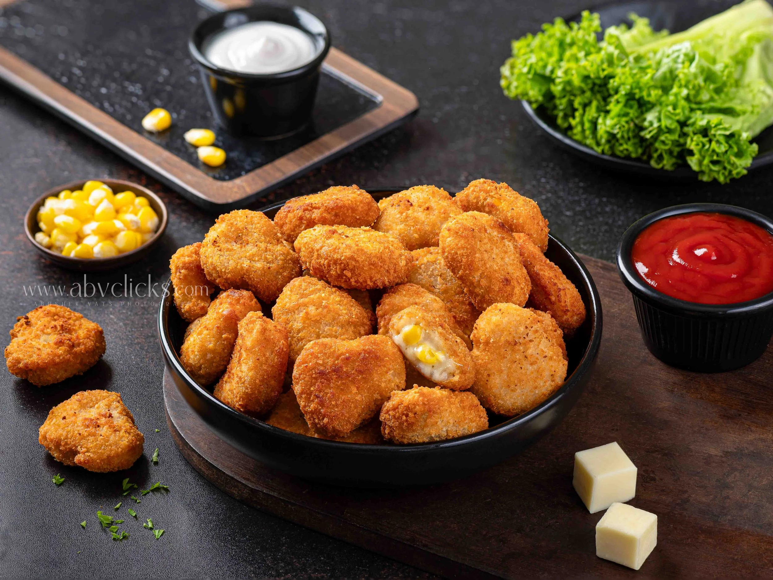 A bowl of fried chicken nuggets with a small bowl of ketchup, a small bowl of corn, a bowl of mixed greens, and a small cup of ranch dressing on a black surface.