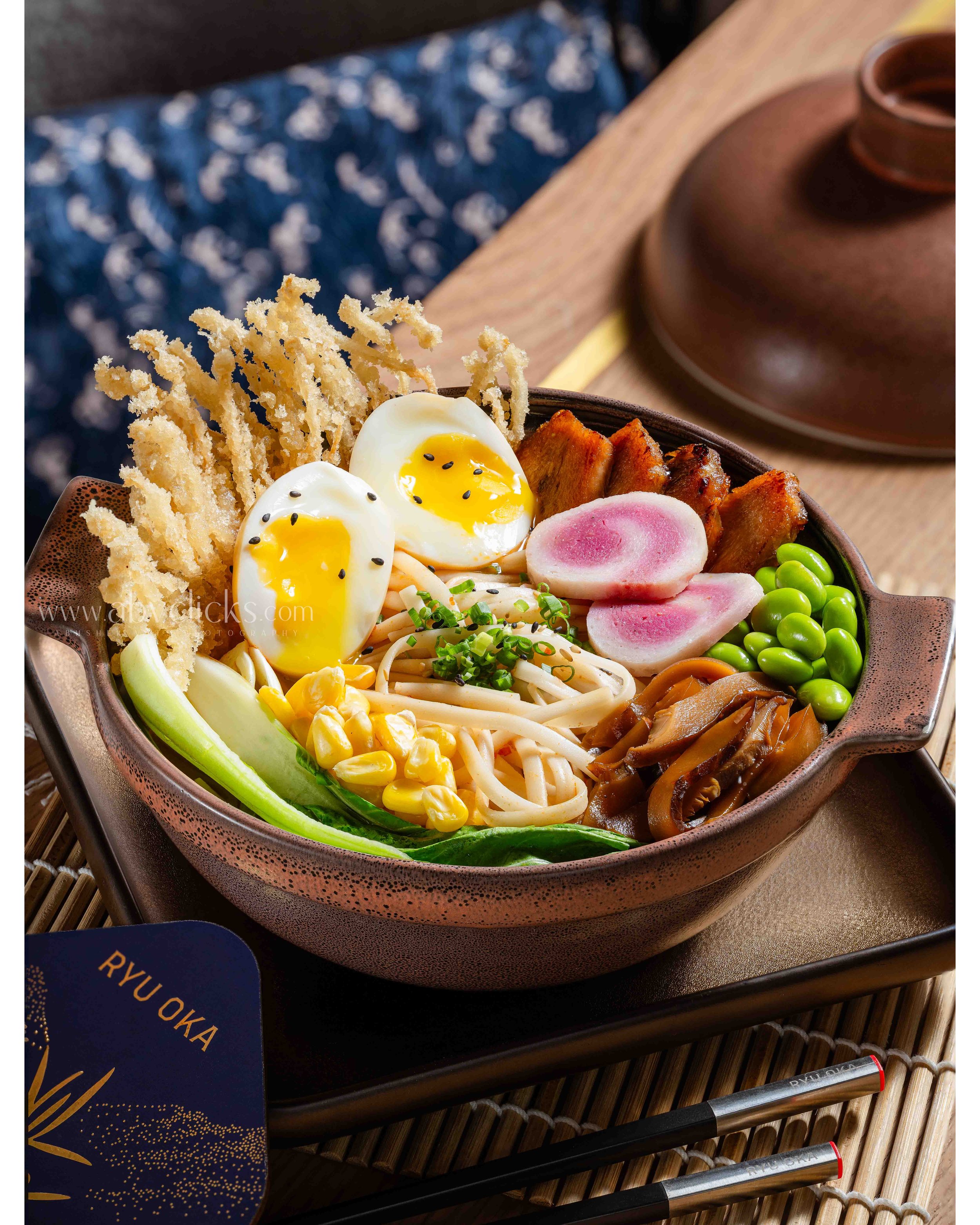 A bowl of ramen with soft-boiled eggs, sliced radish, green peas, corn, grilled meat, and some crispy fried noodles, on a wooden table with chopsticks nearby.