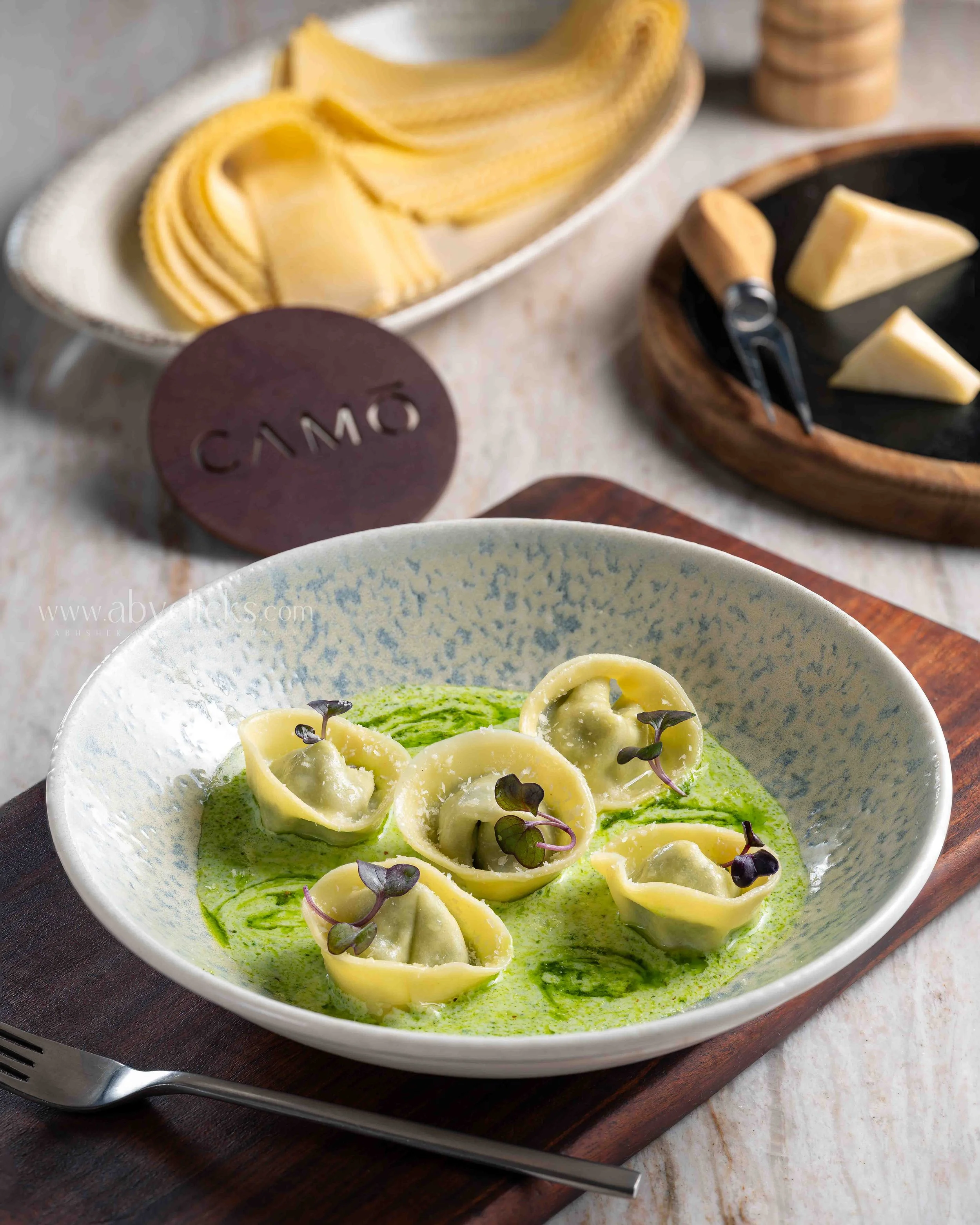 Close-up of a bowl of tortellini pasta with green sauce and microgreens on a dark wooden tray, with a fork beside the bowl.
