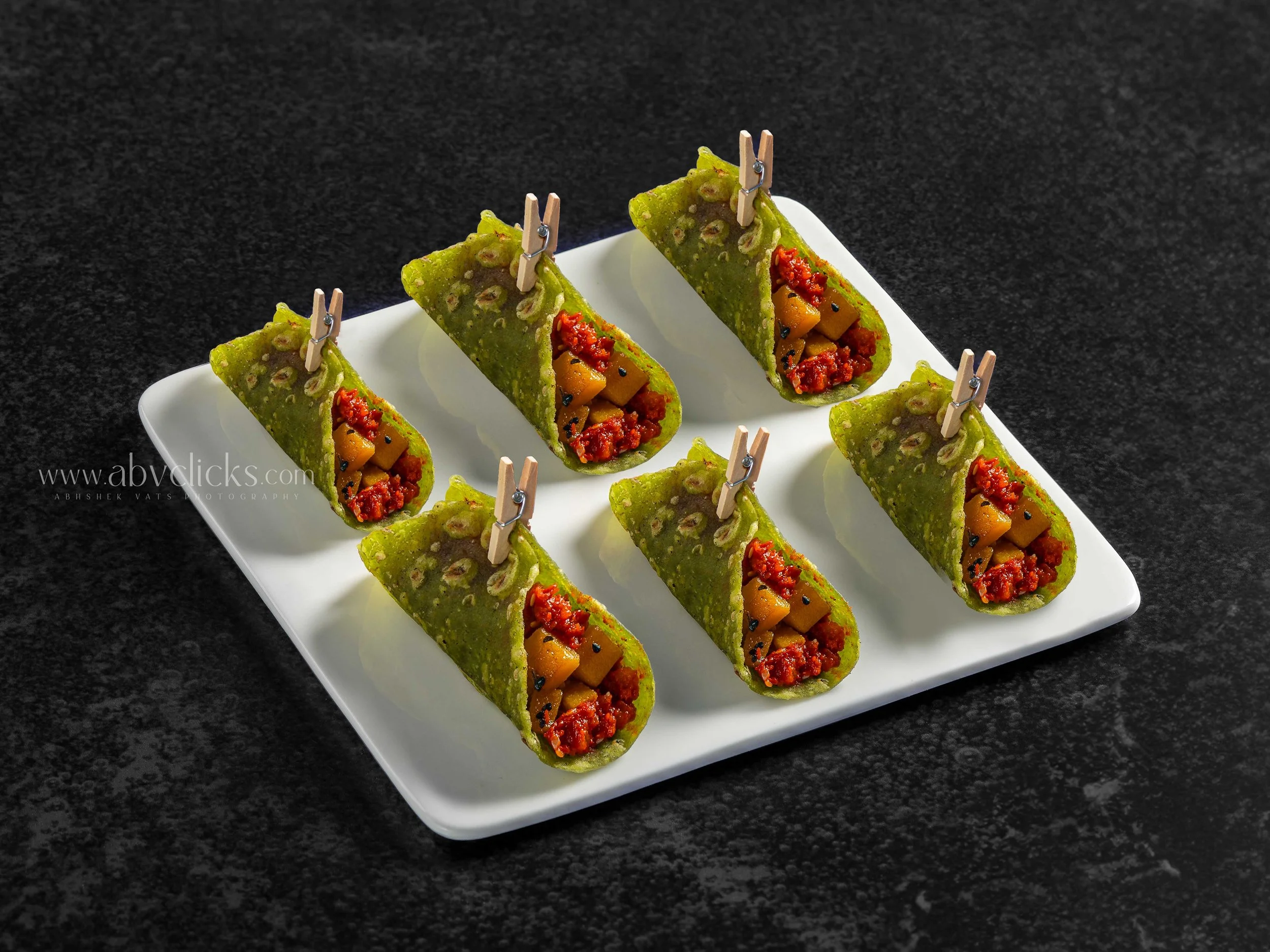 Several green crispy rolls filled with chopped yellow and red vegetables on a white rectangular plate, placed on a black surface.