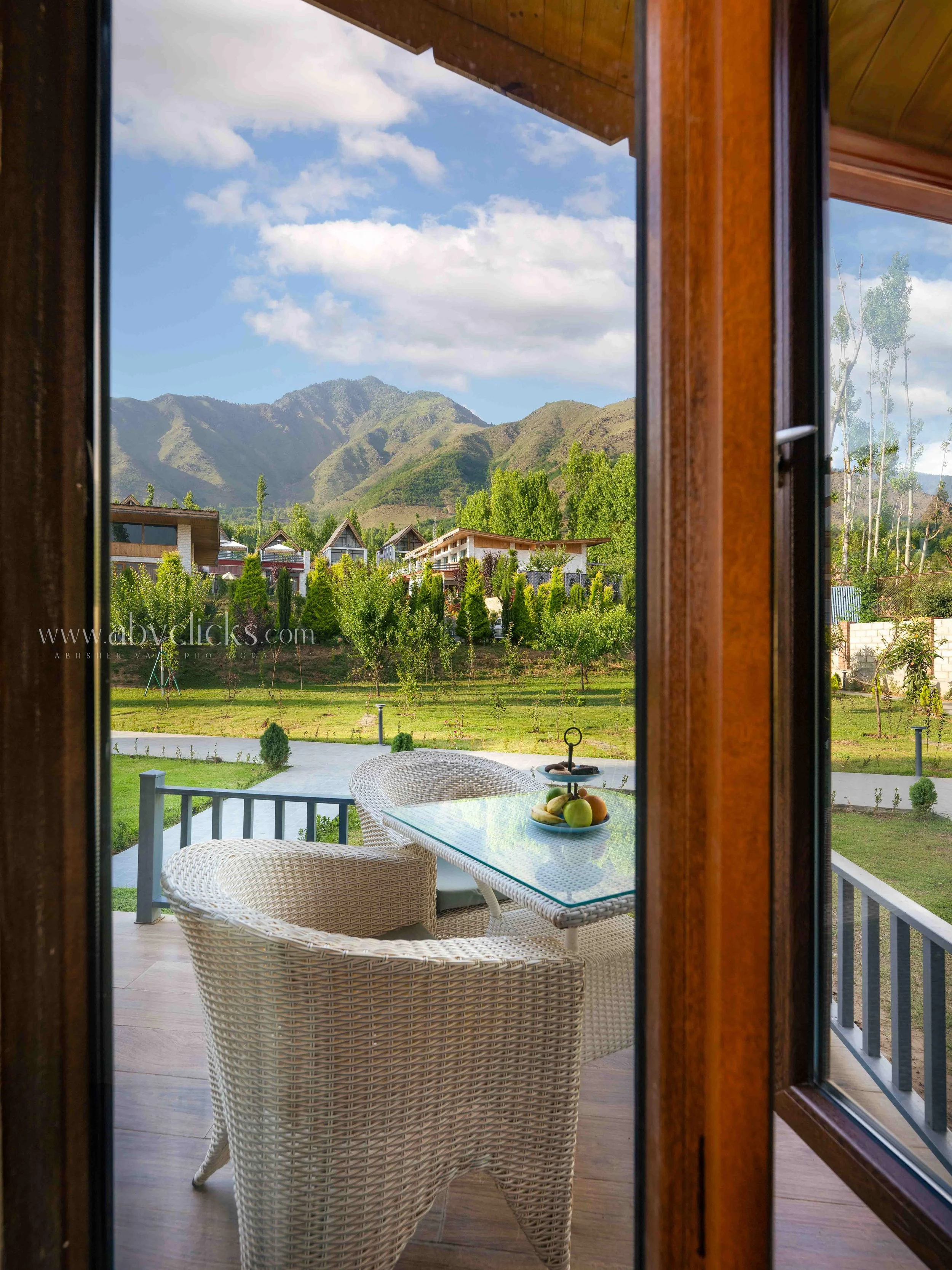 View through a wooden framed window showing a balcony with rattan chairs and a table with a bowl of fruits, overlooking a green garden with trees and a mountain range in the background under a partly cloudy sky.