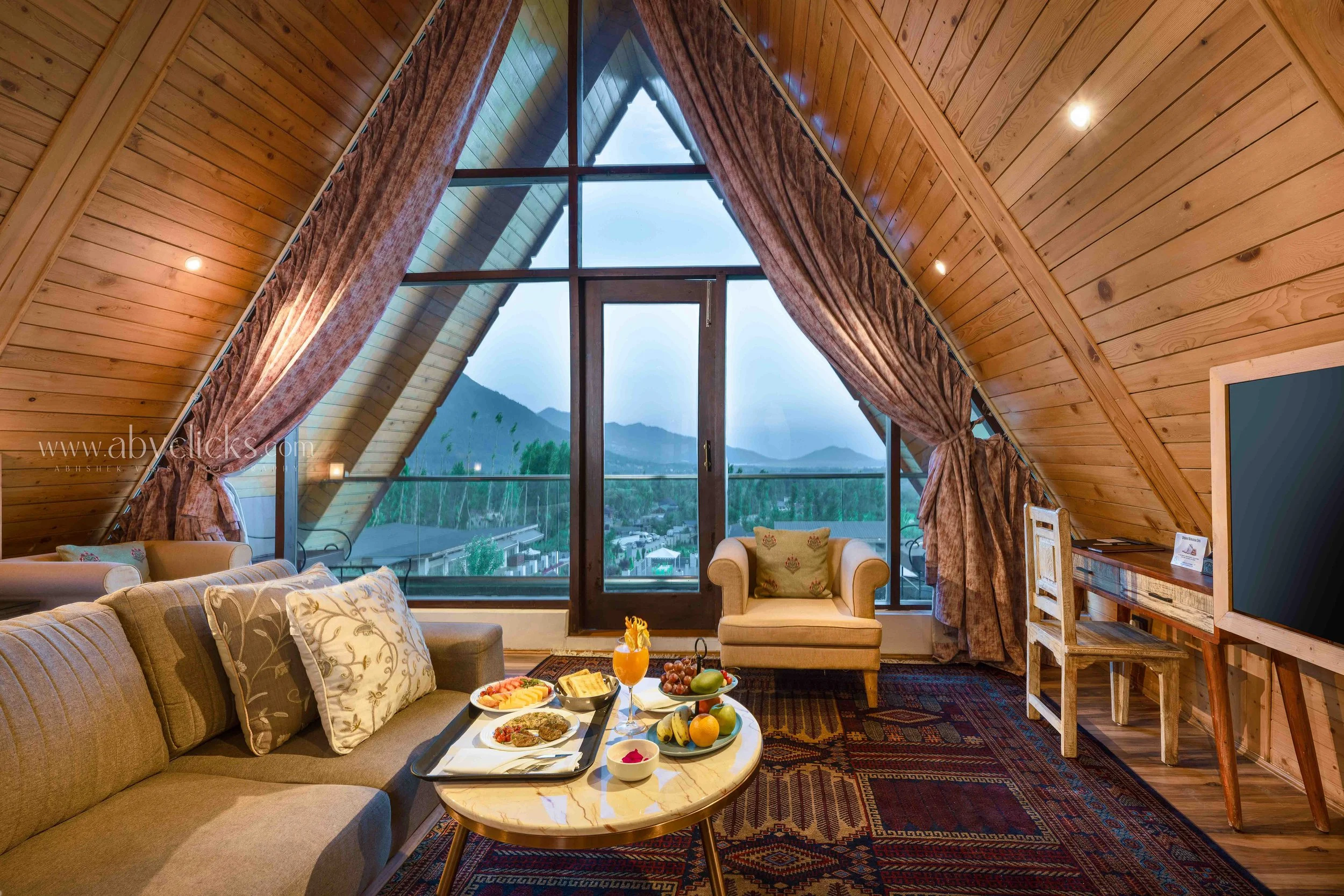 A cozy living room with a large triangular window view of mountains. The room has wooden walls and ceiling, with brown curtains on the window. There is a beige sofa with patterned pillows, a coffee table with breakfast food, and a small armchair. A d