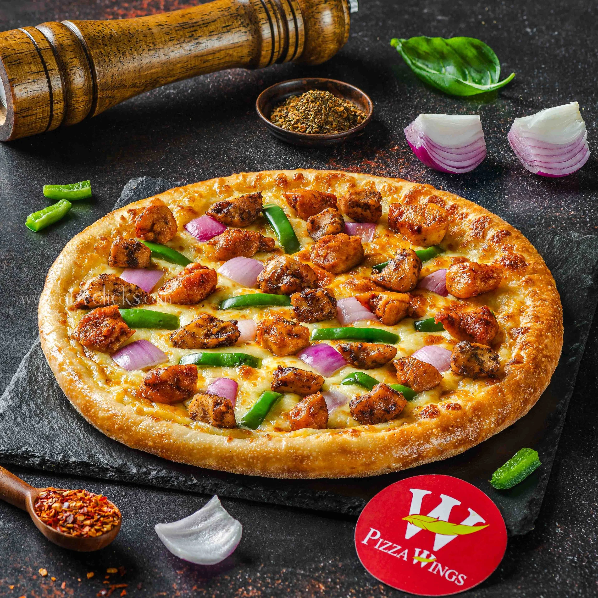 A partially visible pizza with toppings including green peppers and chicken, on a black surface. Surrounding the pizza are a wooden pepper grinder, a small bowl of crushed spice, a green basil leaf, sliced red onion, and a small section of Italian se