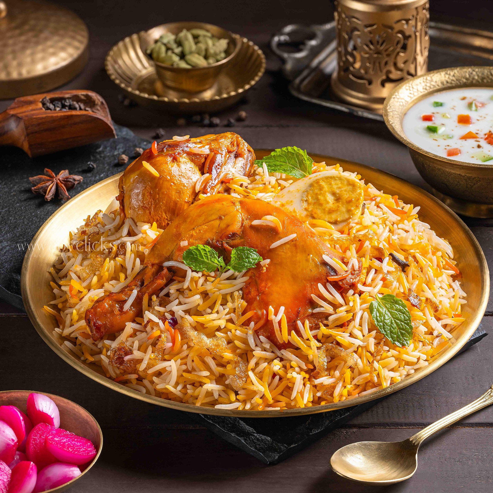 Plate of Indian biryani rice garnished with mint leaves, served with chicken drumsticks. In the background, there are bowls of green cardamom pods, a bowl of chopped vegetables in white sauce, and Indian spices including star anise. The dish is on a 