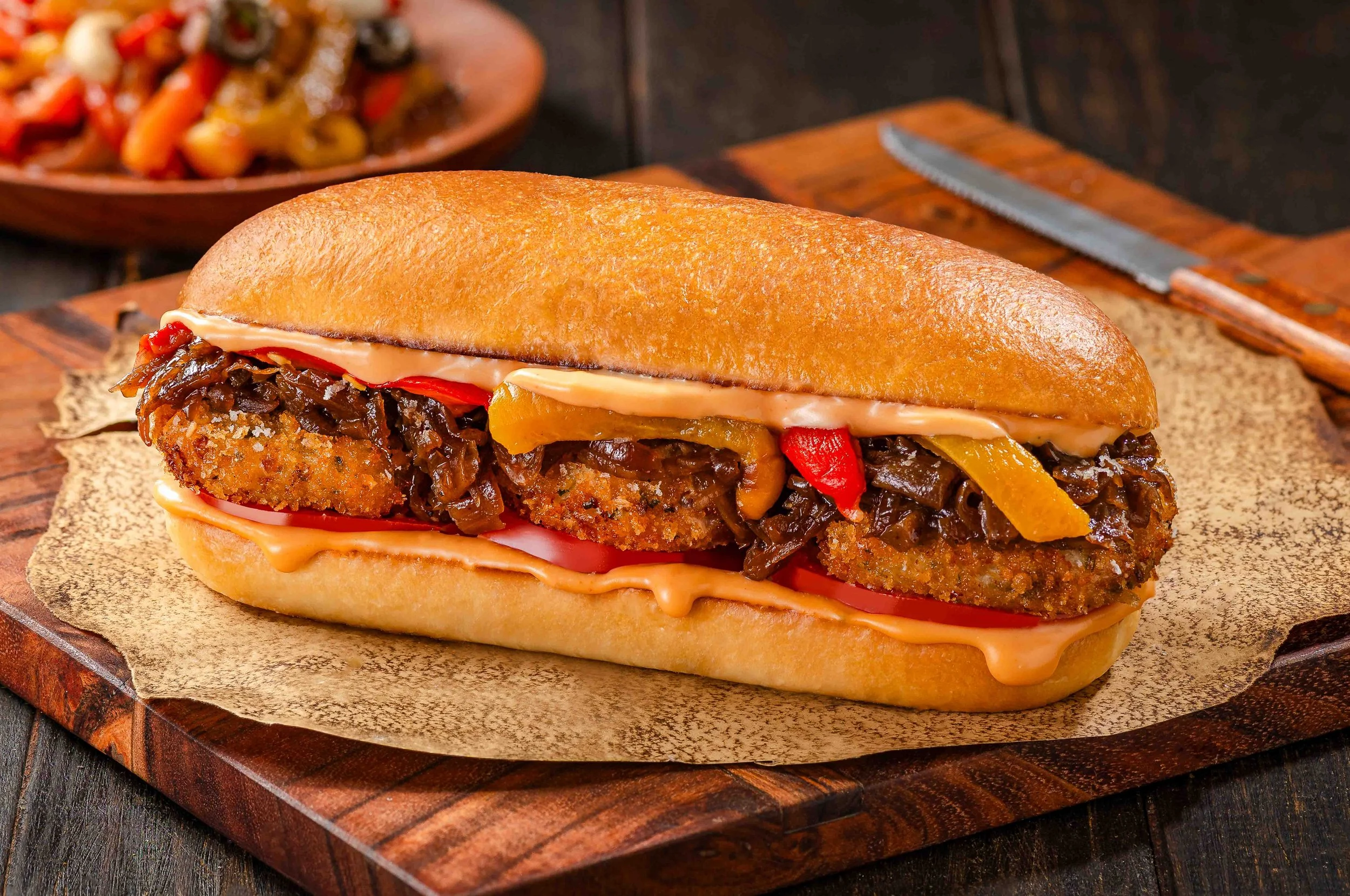 A sandwich filled with shredded barbecue pork, sliced tomatoes, and mayonnaise in a crusty bread bun, placed on a wooden board with a knife in the background and a bowl of grilled vegetables in the blurred background.