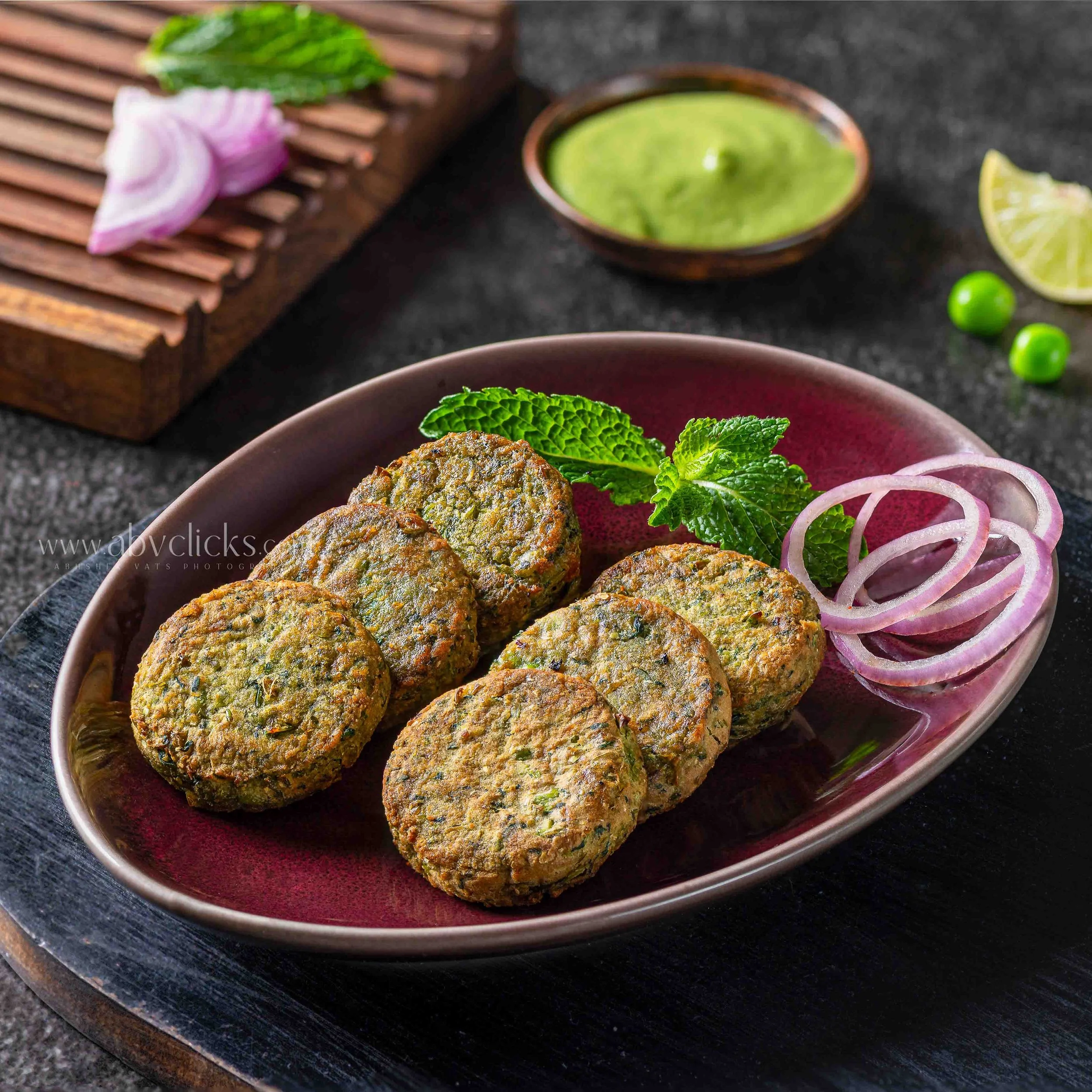 Close-up of falafel balls garnished with mint leaves on a reddish plate, with sliced onions and limes in the background on a black surface.