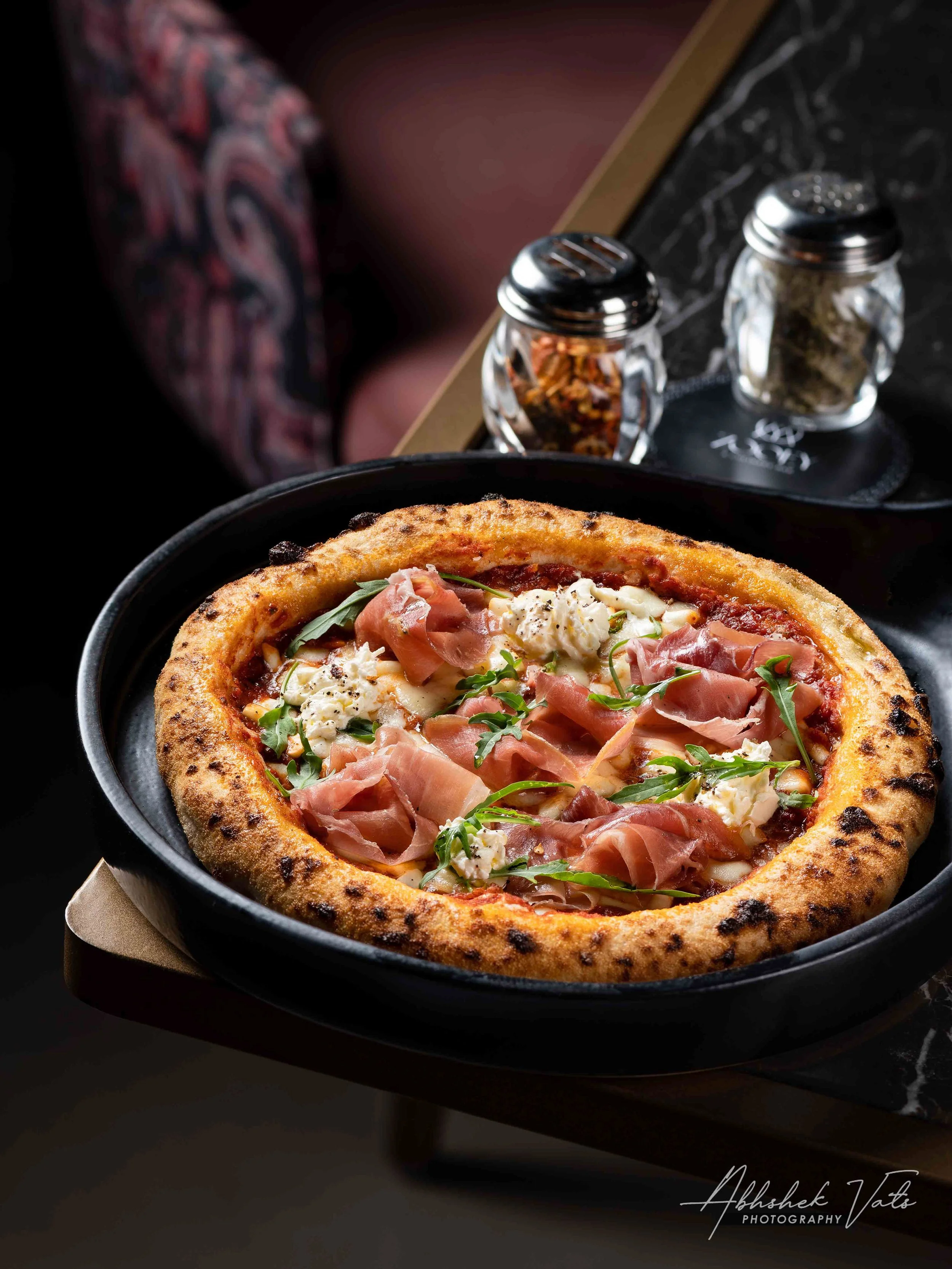 A close-up of a pizza with a golden crust, topped with prosciutto, fresh herbs, and cheese, served in a black dish on a dark surface. In the background, there are two glass jars, one with crushed red pepper and the other with dried herbs, and a black