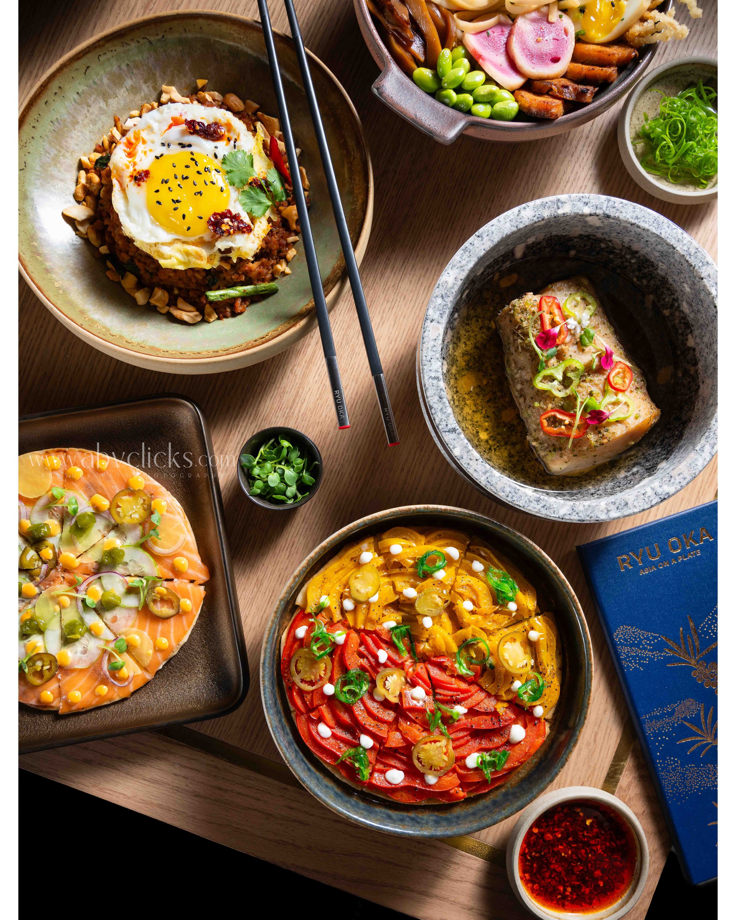 Assorted Asian dishes on a wooden table, including a bowl of rice topped with a fried egg, an assortment of seafood and vegetables, a bowl of salmon sashimi, a spicy fish dish in a stone bowl, and a spicy dipping sauce.