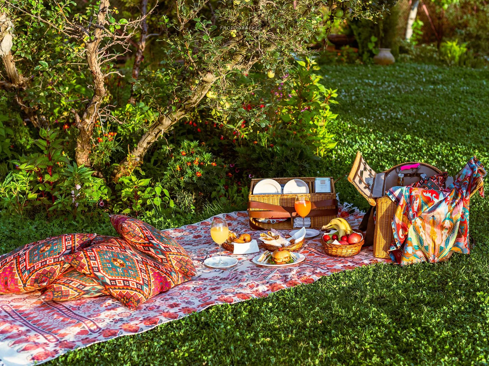 A cozy outdoor picnic setup on a floral blanket in a lush green garden with trees and bushes. The scene includes colorful patterned pillows, a wicker picnic basket with plates and utensils, and a variety of food and drinks including glasses of orange