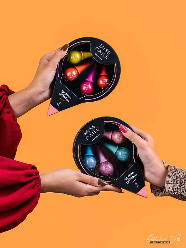 Two hands holding black circular packages of Miss Nails ice cream collection, each containing colorful nail polish bottles shaped like ice cream cones, against an orange background.
