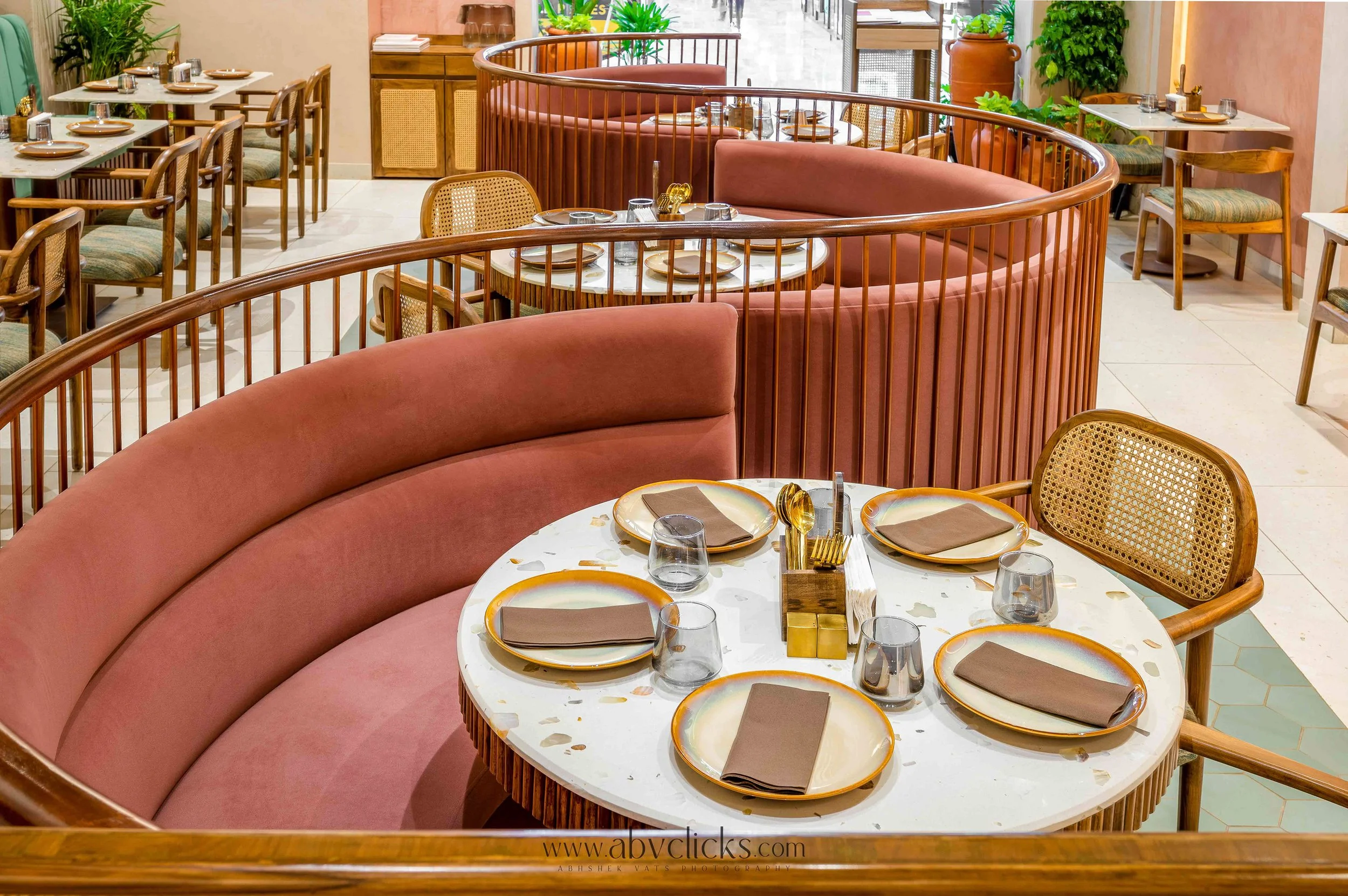 A restaurant dining area with round marble-top tables set with gold-rimmed plates, brown napkins, glasses, and gold utensils, surrounded by pink curved booths and wooden chairs with woven cane backs. Green plants are visible in the background.