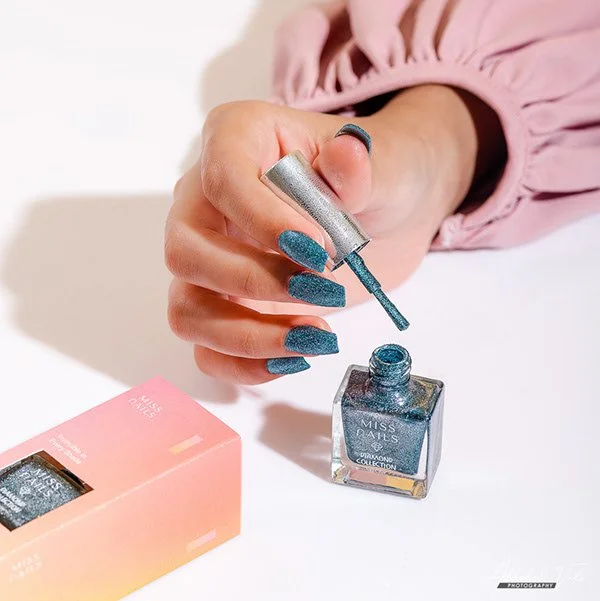 Person with blue, textured nails applying blue nail polish from a small brush, with a pink box beside on a white surface.